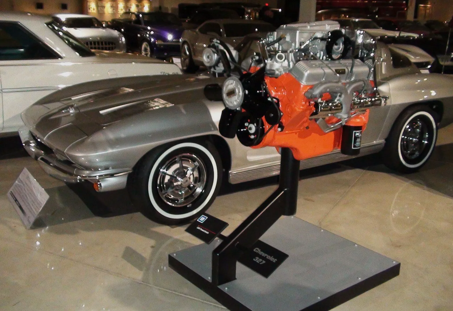 A silver Corvette Sting Ray with whitewall tires on display indoors, with a fuel-injected Chevrolet 327 V8 with an orange block standing on its own display stand in front of the car.