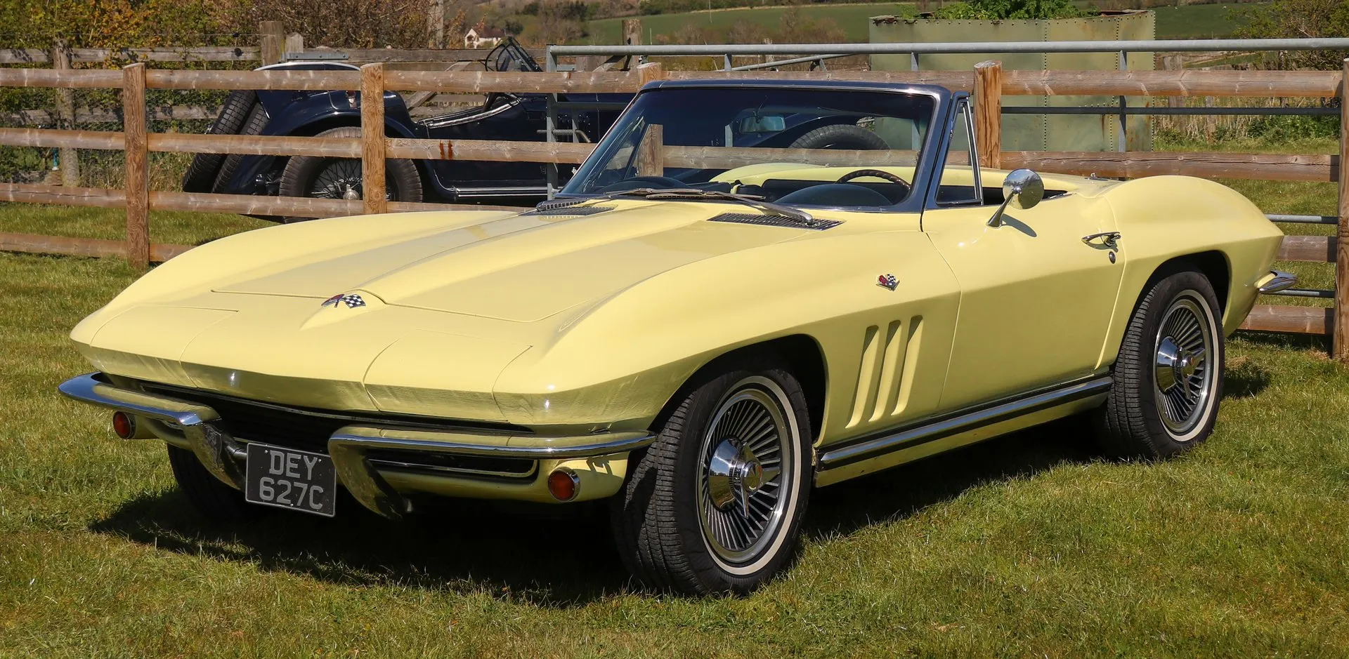 A pale yellow Corvette convertible with its top down, parked on grass beside a wooden fence and seen from the front left, with a flat hood, three vertical fender vents, whitewall tires and a British registration plate.
