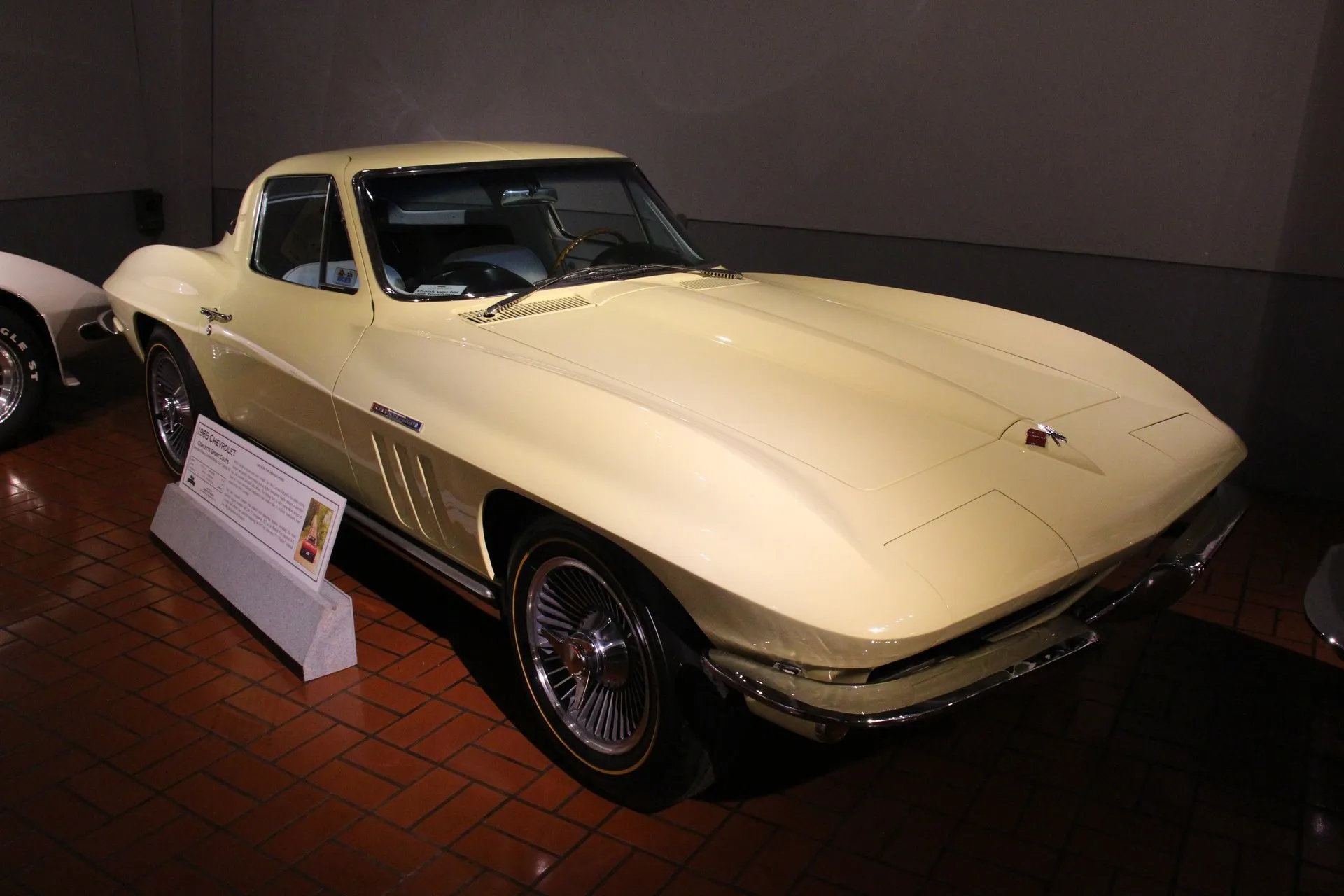 A pale yellow 1965 Corvette coupe on a brick floor in a museum, seen from the front right, with a flat hood, three vertical vents in the front fender, spoked wheel covers, gold-stripe tires and a placard on a stand beside the car.