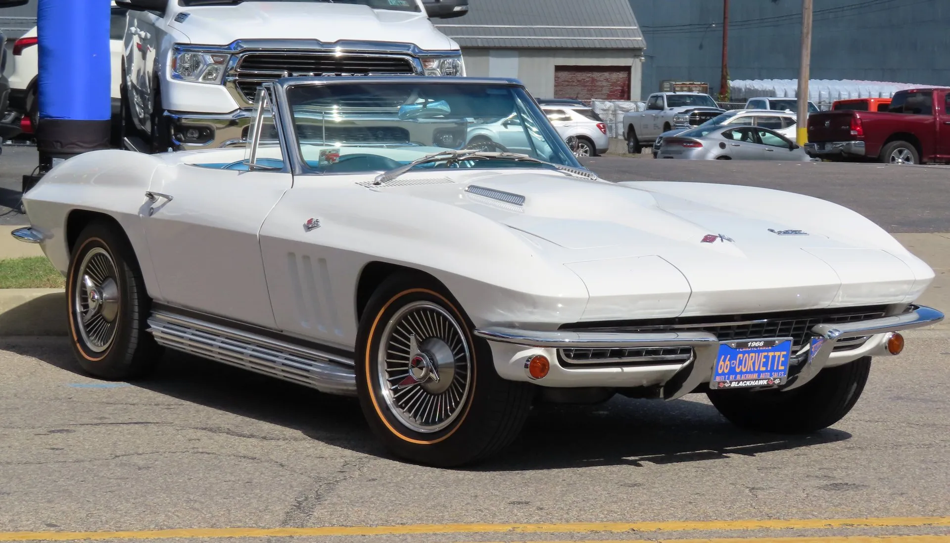 A white 1966 Corvette convertible with its top down, seen from the front right on a dealer lot, with a raised hood center, three fender vents, chrome side exhaust covers, gold-stripe tires and a front plate reading 66 CORVETTE.
