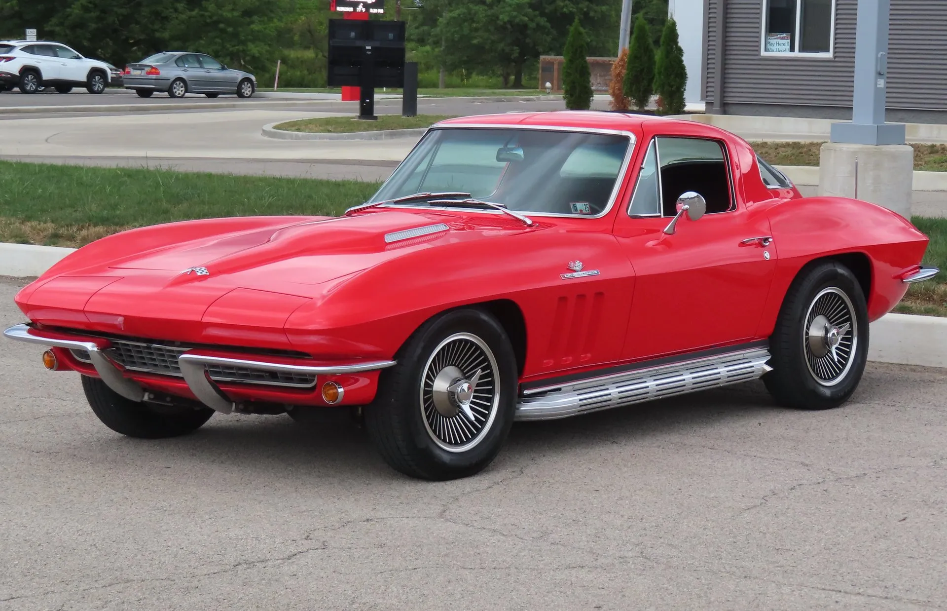 A red 1966 Corvette Sting Ray coupe in a parking lot, seen from the front left, with a raised center section on the hood, three vertical vents and a crossed-flags emblem on the front fender, chrome side exhaust covers below the door and wheel covers with spinner hubs.