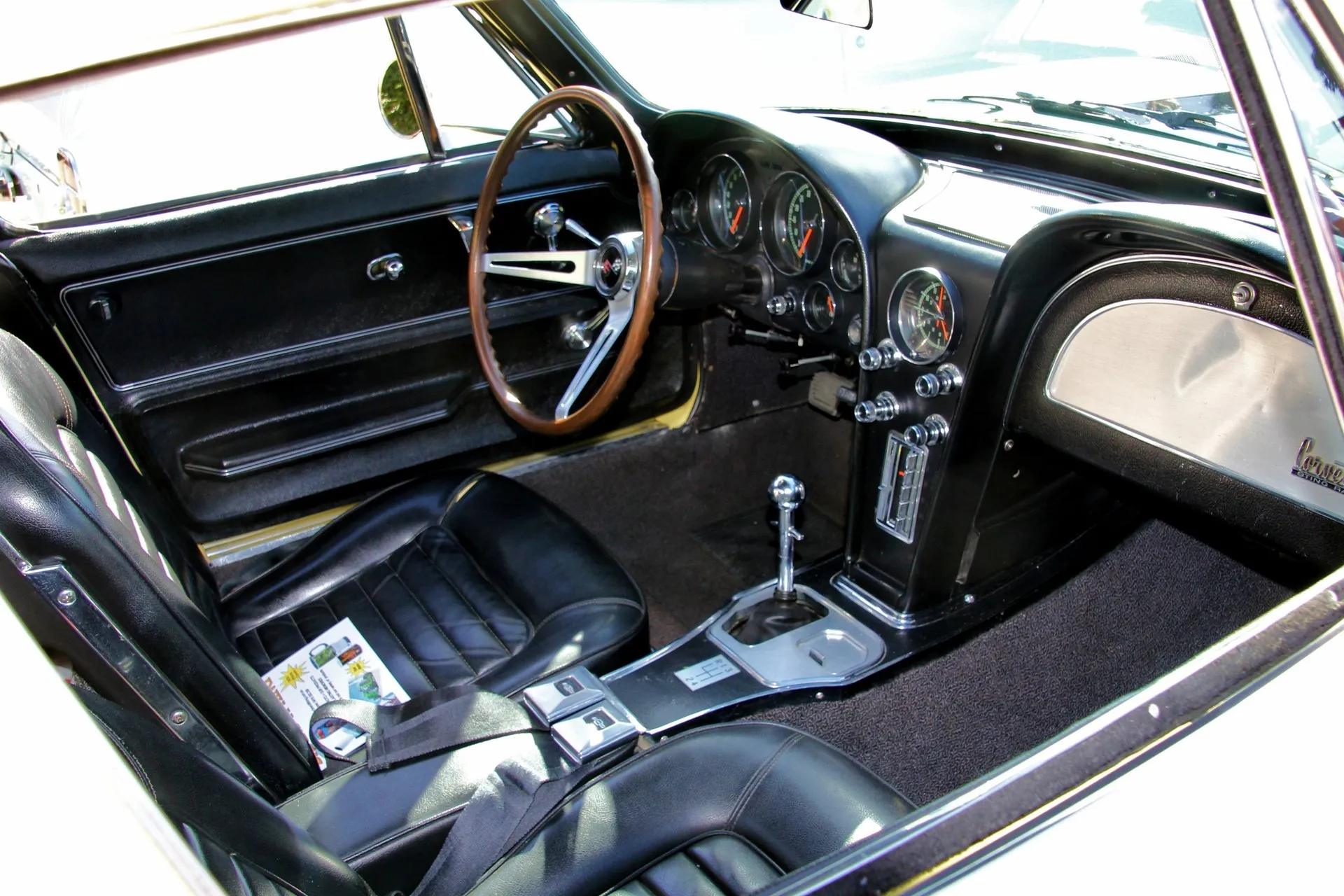 The black interior of a Corvette coupe seen through the open passenger door: pleated seats, a wood-rim steering wheel, a twin-cowl dashboard with round gauges, a four-speed shifter on the center console and a Corvette Sting Ray script on the glove box door.