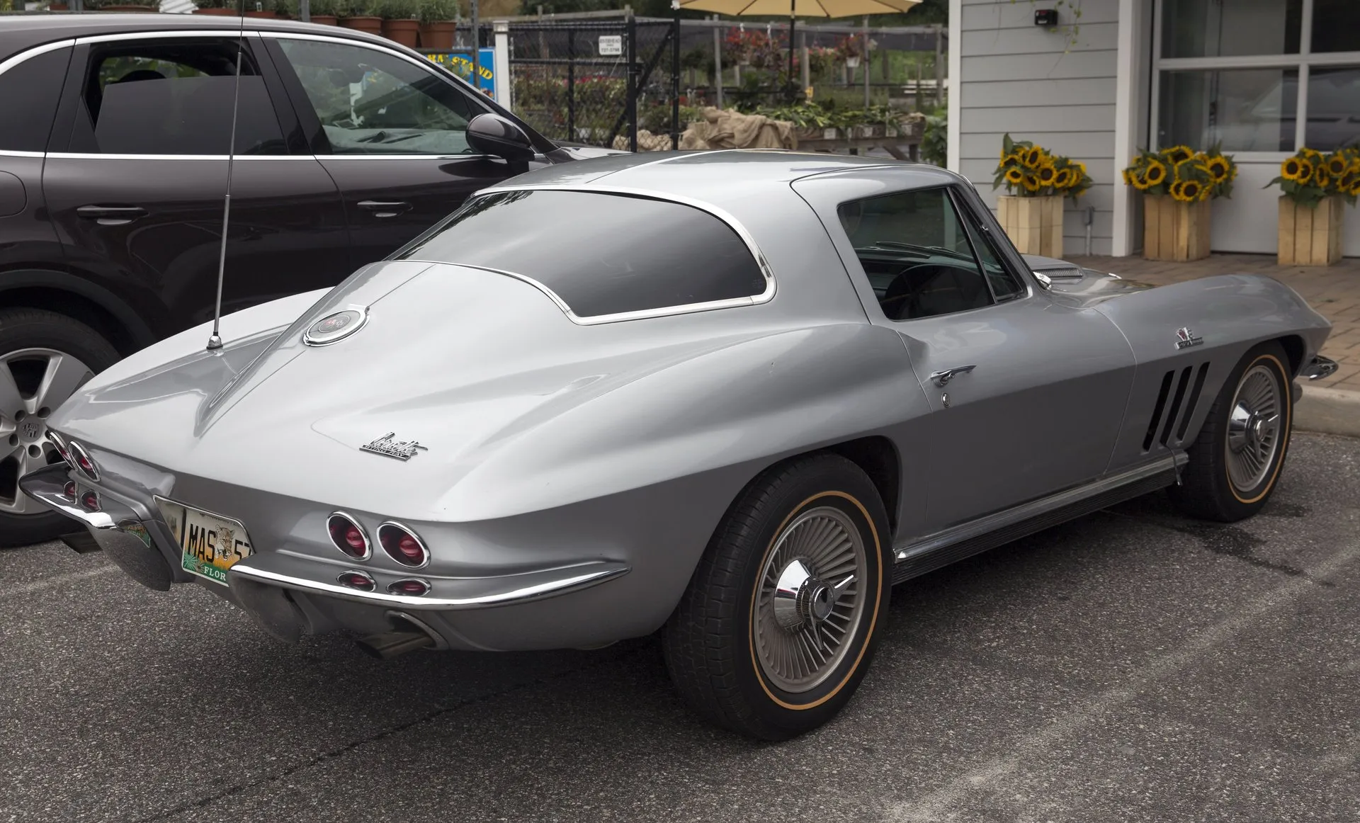 A silver 1966 Corvette coupe in a parking lot, seen from the rear right, with a one-piece rear window, no vent in the roof pillar behind the door, three vertical vents in the front fender, twin round taillights on each side and gold-stripe tires.