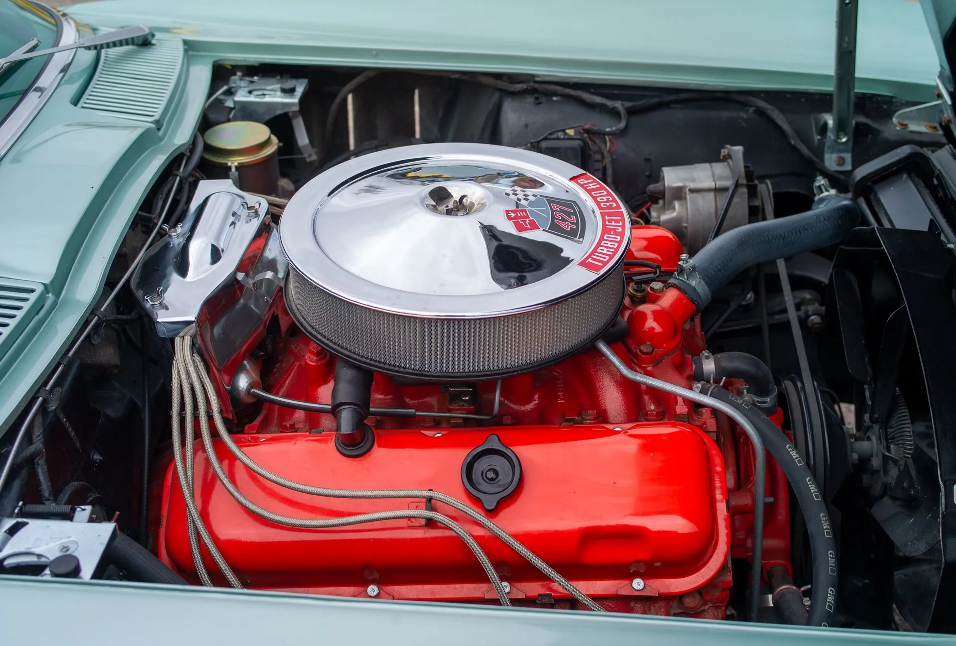 The engine bay of a Corvette with the hood up: an orange big-block V8 under a chrome air cleaner whose decal reads 427 and Turbo-Jet 390 HP, with braided covers on the spark plug wires.