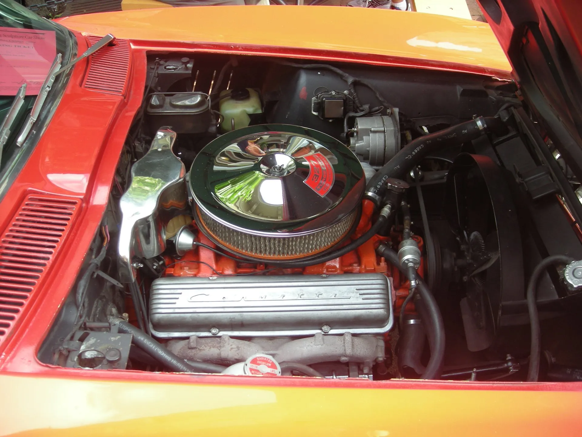The engine bay of a red Corvette seen from the side: an orange small-block V8 under a round chrome air cleaner with a red 327 Turbo-Fire 300 horsepower decal, and a finned aluminum valve cover with Corvette script.