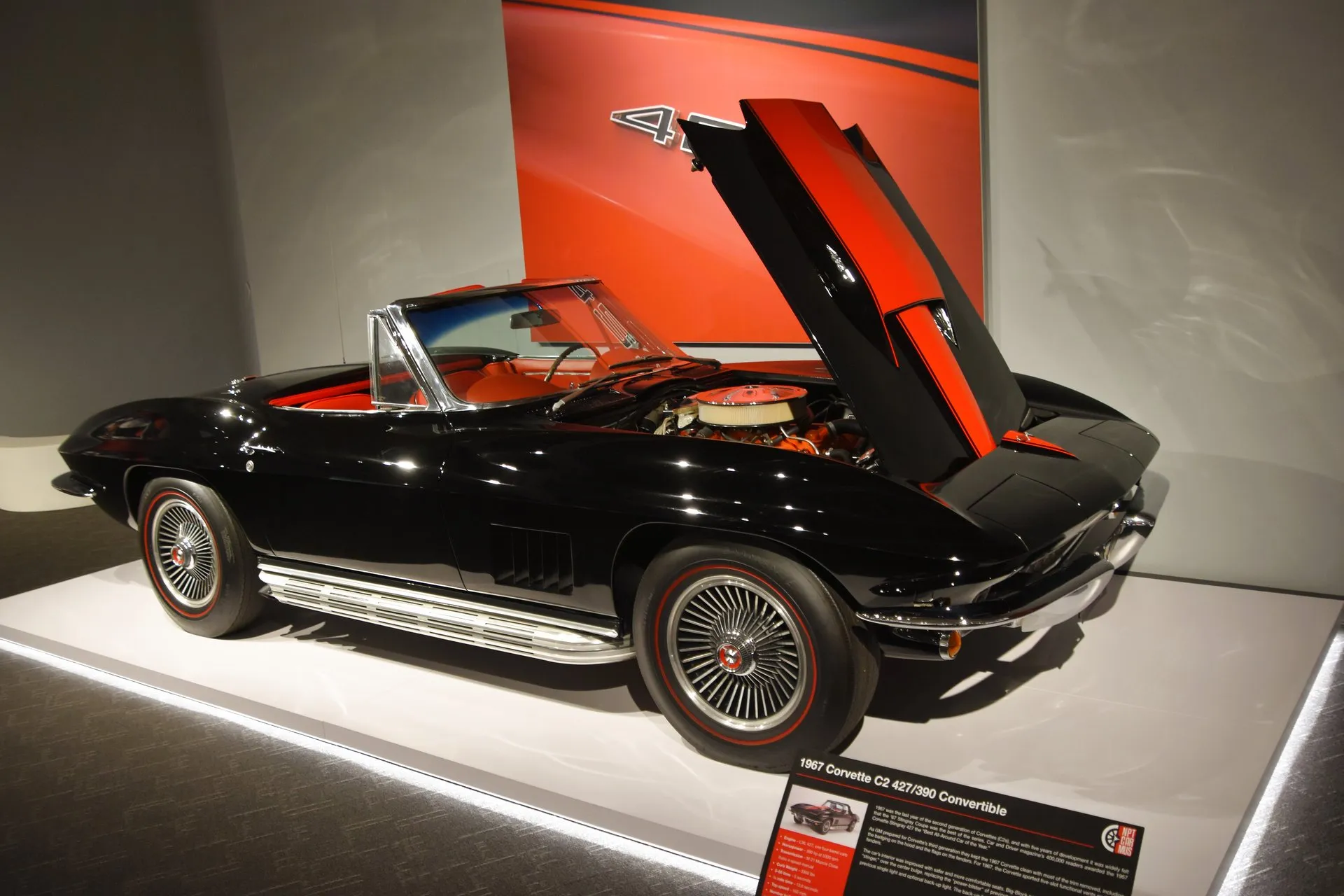 A black 1967 Corvette convertible with a red interior on a museum display, seen from the front right with its hood raised to show a red stripe and a 427 V8 under a round air cleaner, beside a placard reading 1967 Corvette C2 427/390 Convertible.