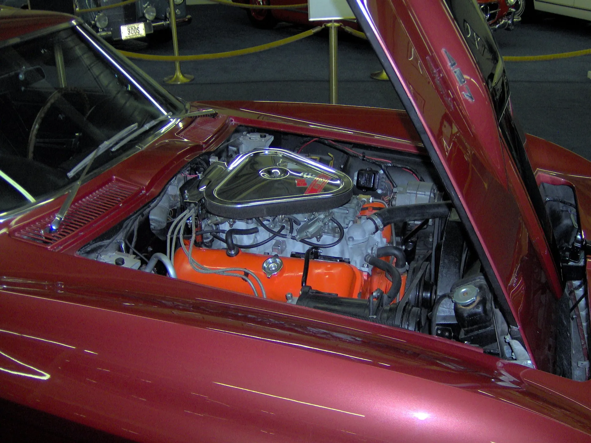 The engine bay of a maroon 1967 Corvette coupe with the hood raised: an orange 427 V8 under a triangular chrome air cleaner that covers three carburetors, in a museum with rope barriers behind the car.