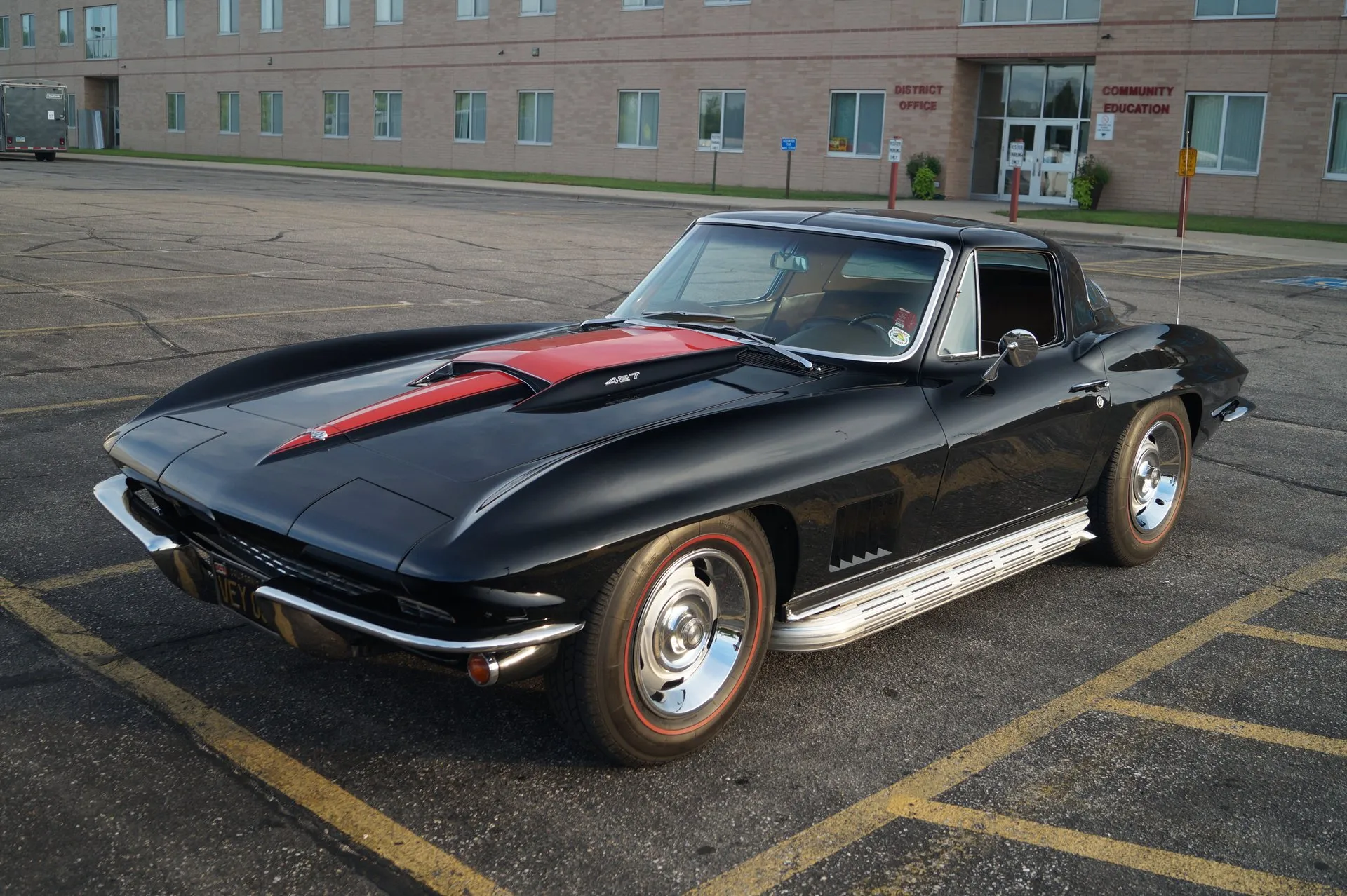 A black 1967 Corvette coupe in a parking lot, seen from the front left, with a red stripe and a 427 call-out on the hood's raised scoop, five angled vents in the front fender, chrome side exhaust covers and Rally wheels with trim rings on red-stripe tires.