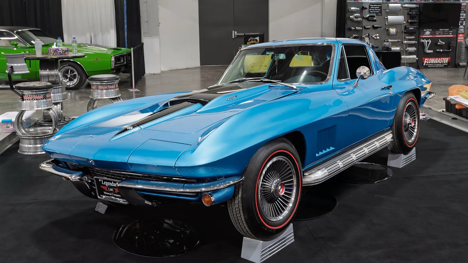 A bright blue 1967 Corvette coupe on a display stand at an indoor show, seen from the front left, with a black stripe on the raised center of the hood, five angled vents in the front fender, chrome side exhaust covers below the door and finned aluminum wheels on red-stripe tires.