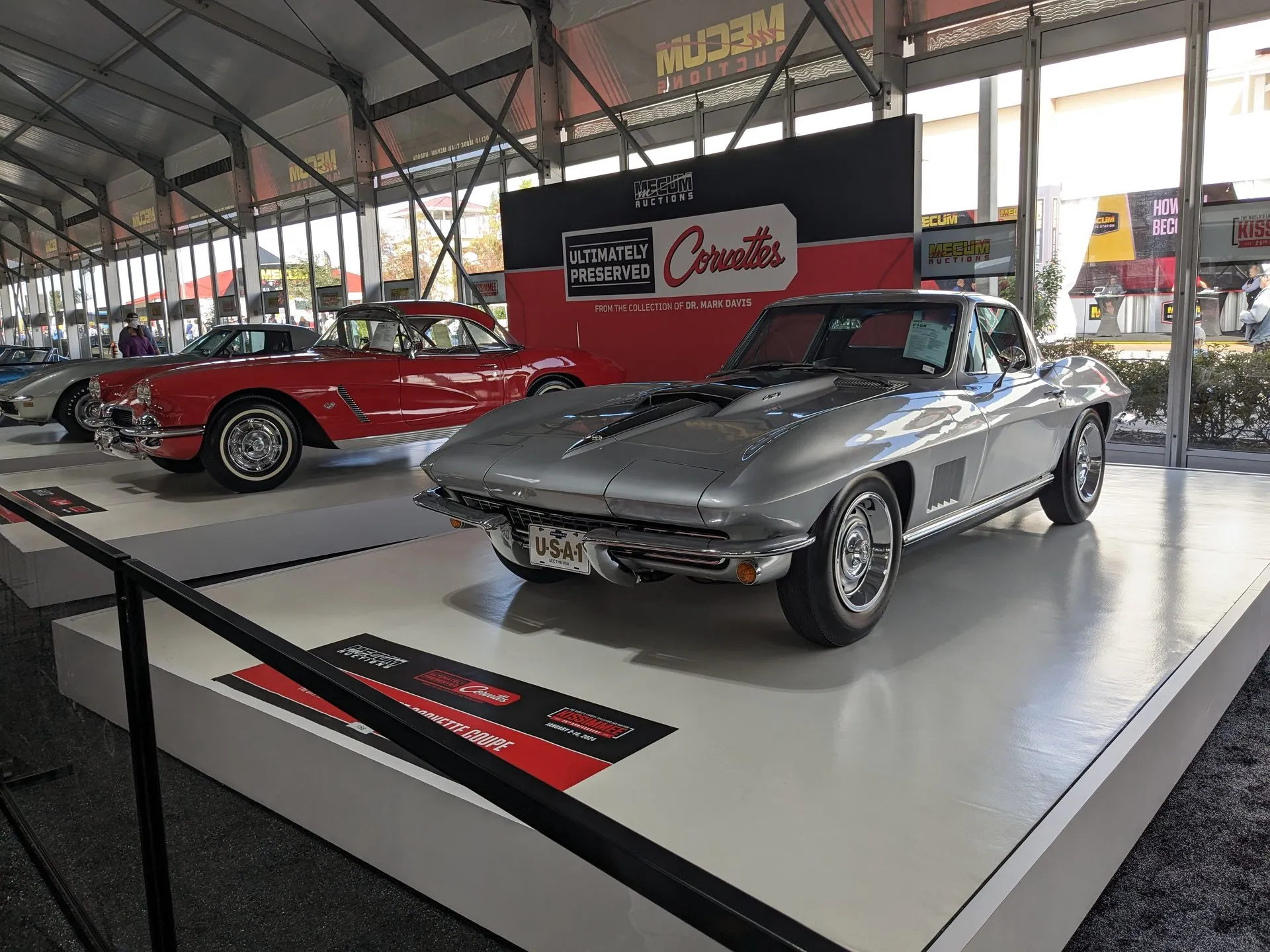 A silver 1967 Corvette coupe on an auction display stand, seen from the front left, with a big-block hood scoop, five vents in the front fender and Rally wheels; a red C1 Corvette is parked behind it.