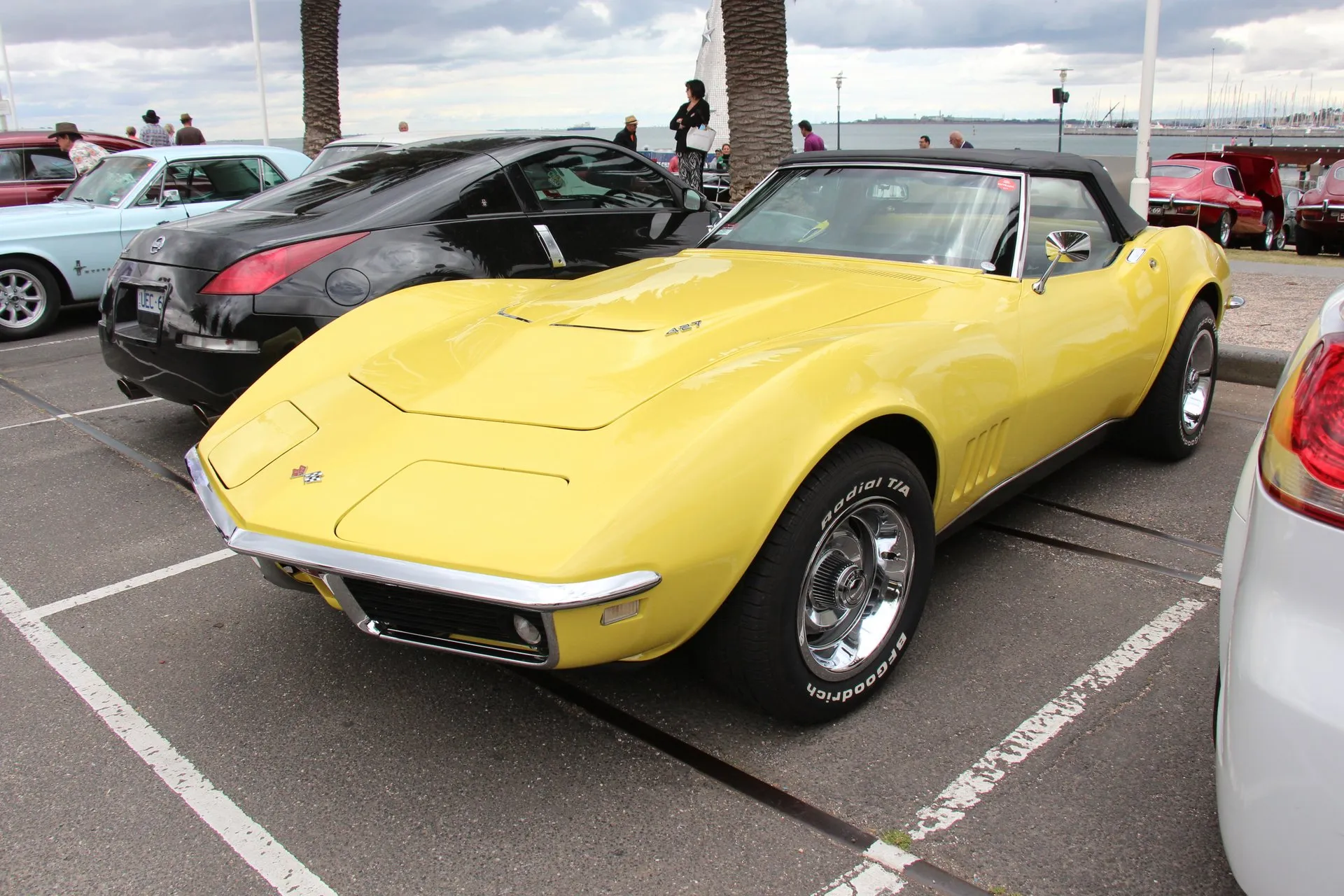 A yellow 1968 Corvette convertible with its black soft top up, parked in a seaside car park and seen from the front left, with a 427 emblem on the raised center of the hood and white-lettered radial tires.