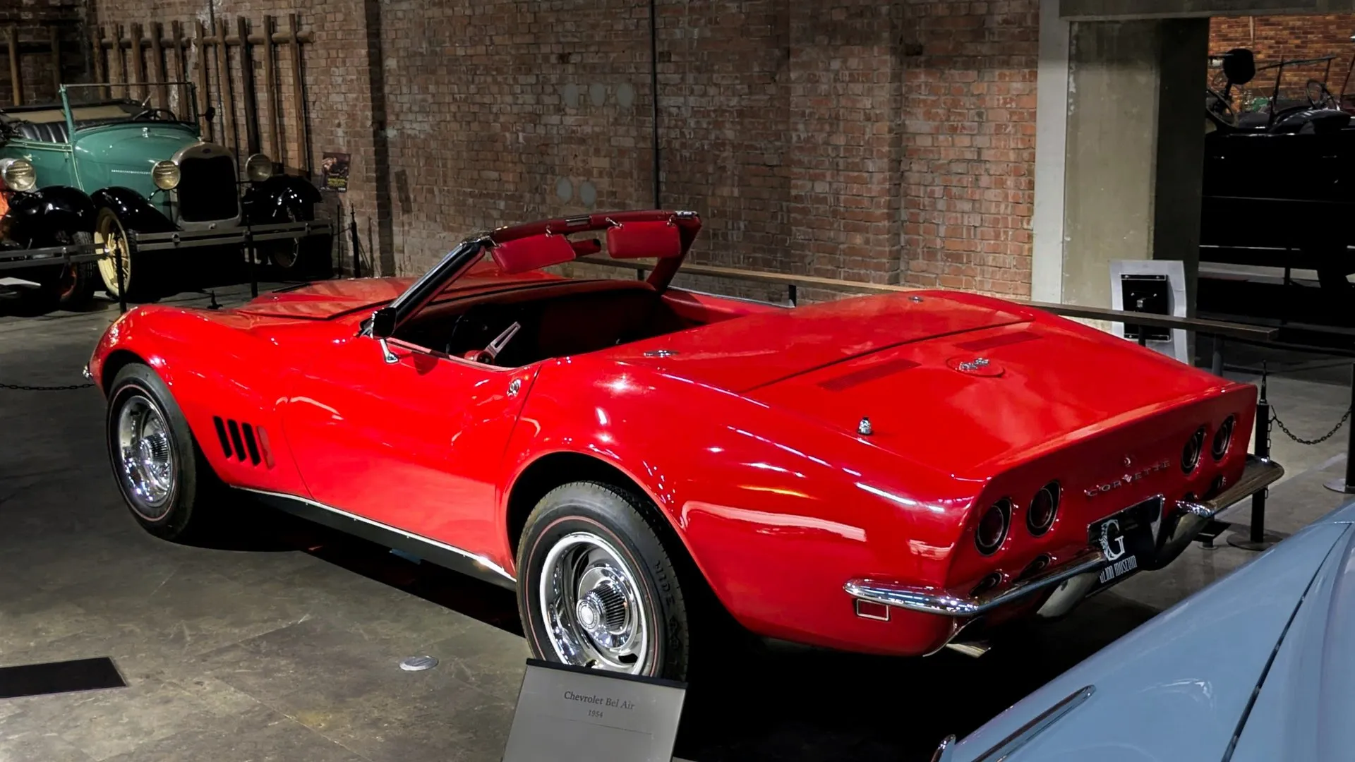 A red 1968 Corvette convertible with its top down in a museum hall, seen from behind and to the left, with four round taillights, a Corvette nameplate between them and a small back-up lamp in the panel below the chrome rear bumper.
