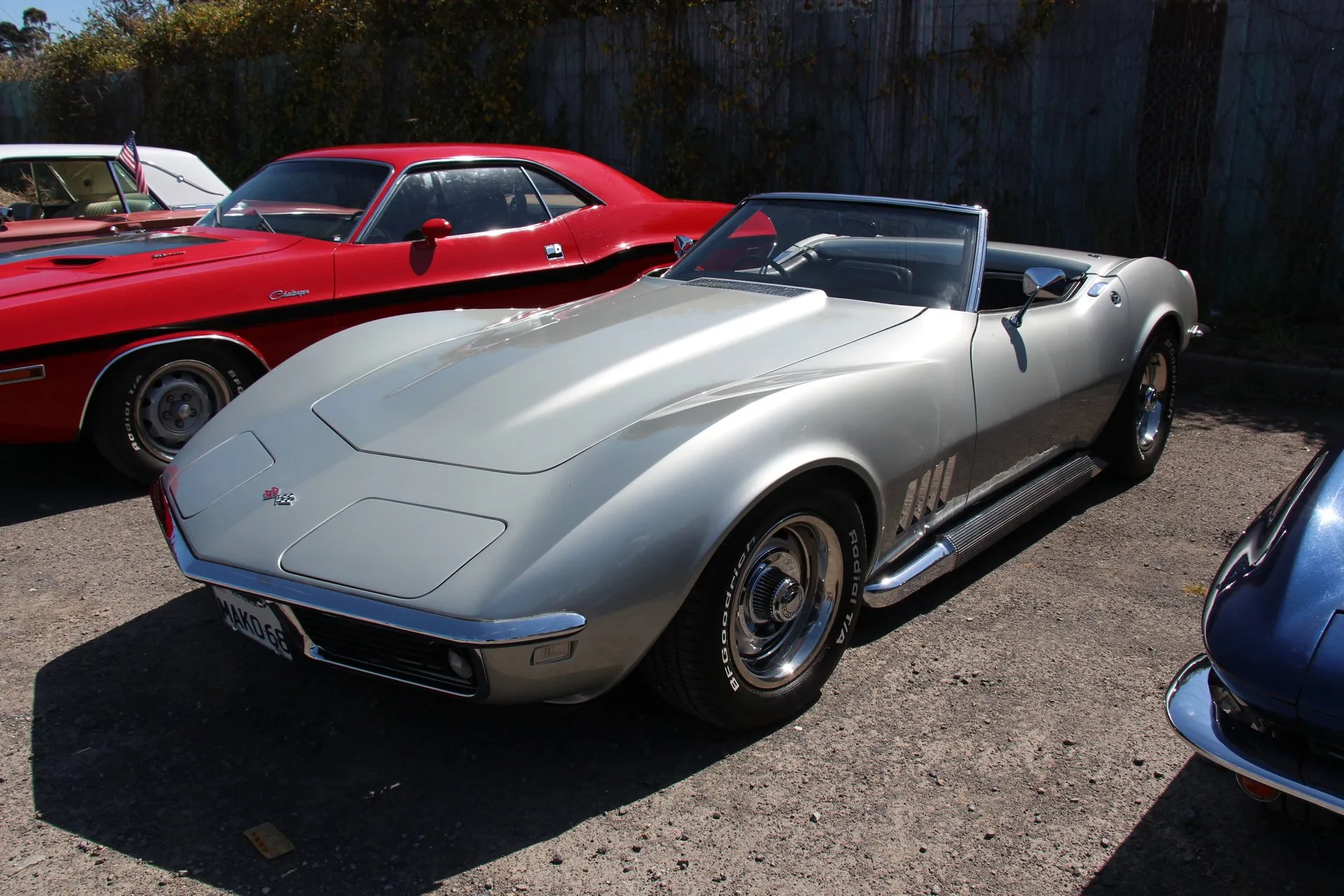 A silver 1968 Corvette convertible with its top down, parked on gravel and seen from the front left, with chrome side exhaust pipes running under the doors and white-lettered radial tires.