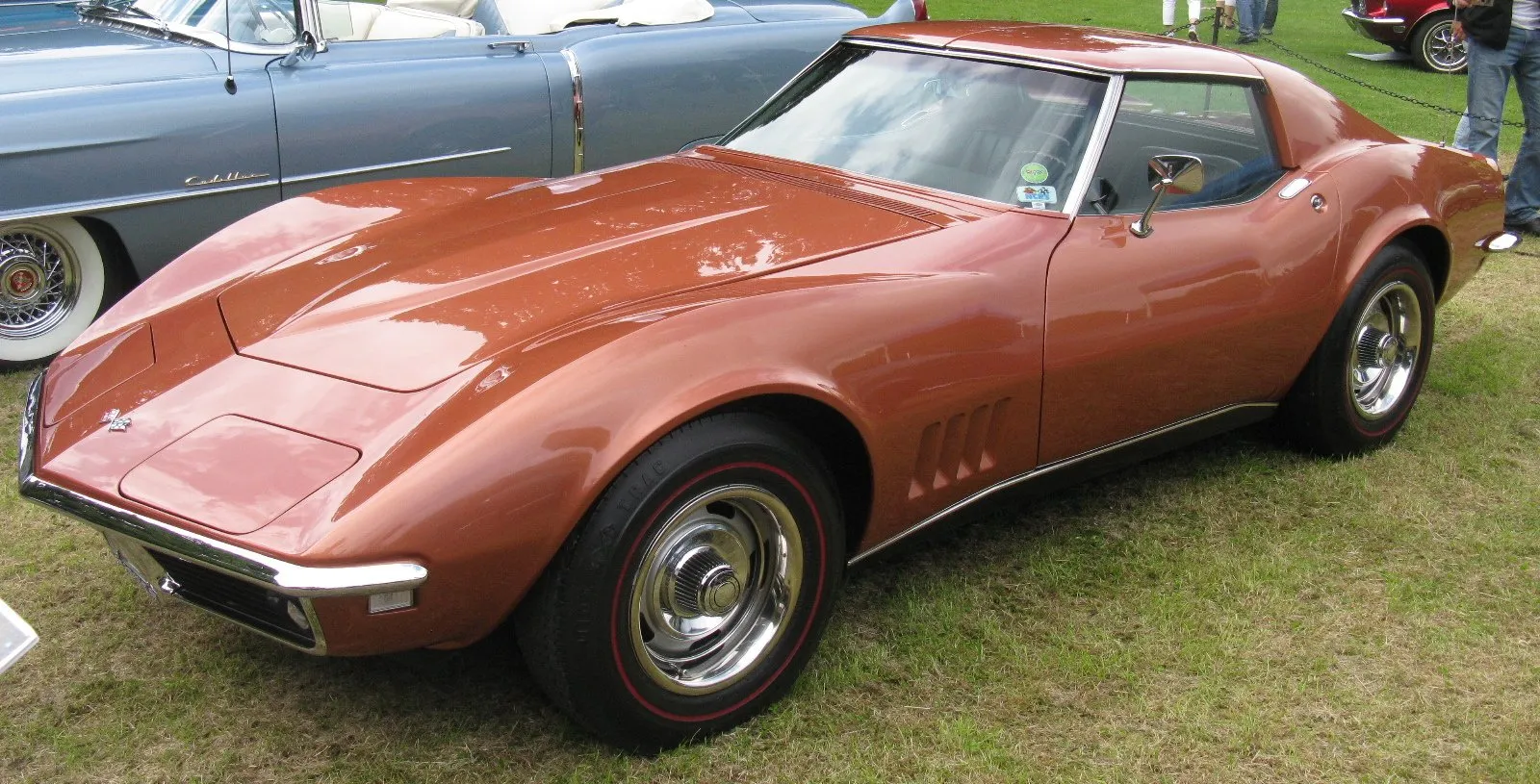 A copper-bronze 1968 Corvette coupe parked on grass at a car show, seen from the front left, with its hidden headlamps closed, four vertical slots behind the front wheel and no script above them, chrome wheel trim rings and red-stripe tires.