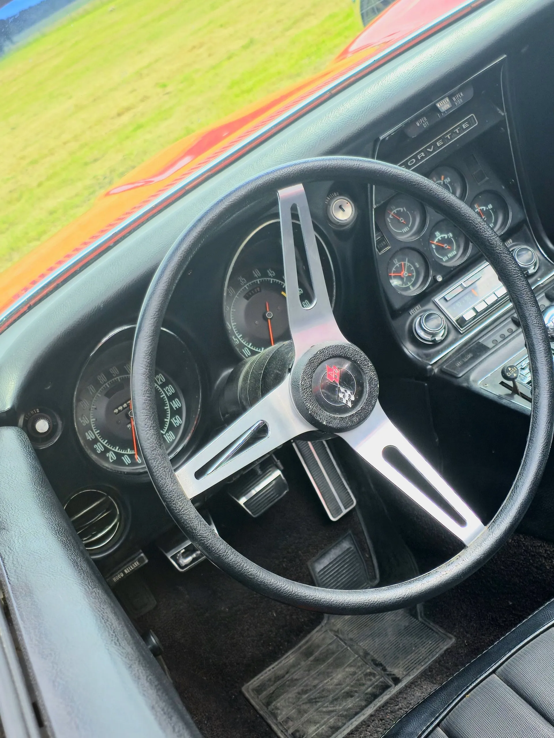 The driver's side of a C3 Corvette cabin: a three-spoke steering wheel with a black rim, metal spokes and a crossed-flags horn button, a speedometer to 160 mph and a tachometer behind it, a key switch marked IGNITION in the dash beside the column, and a center stack of small gauges under a Corvette nameplate.