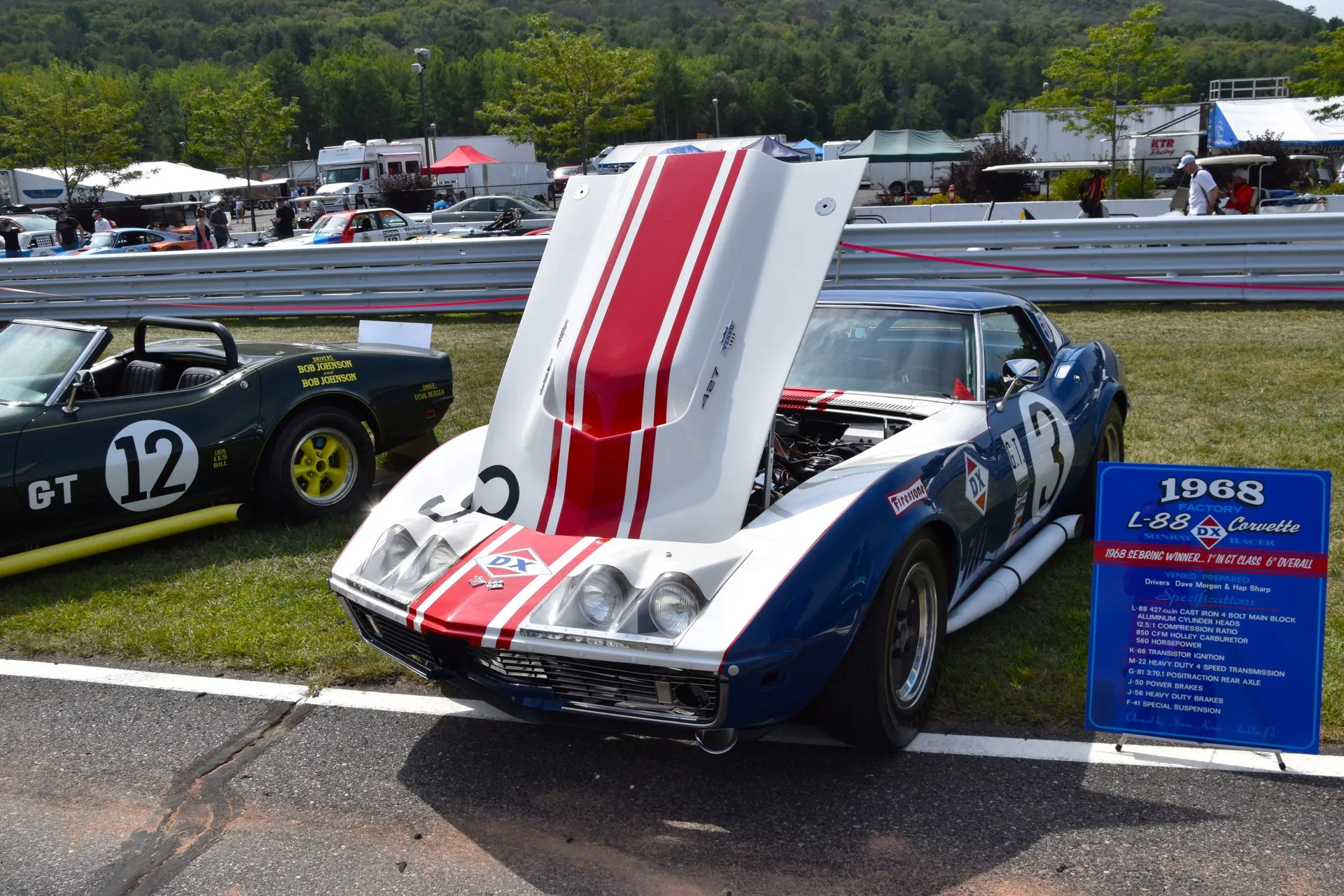 A white, red and blue race-liveried C3 Corvette coupe with its hood raised on the grass at a race track, beside a blue display placard headed 1968 Factory L-88 Corvette.