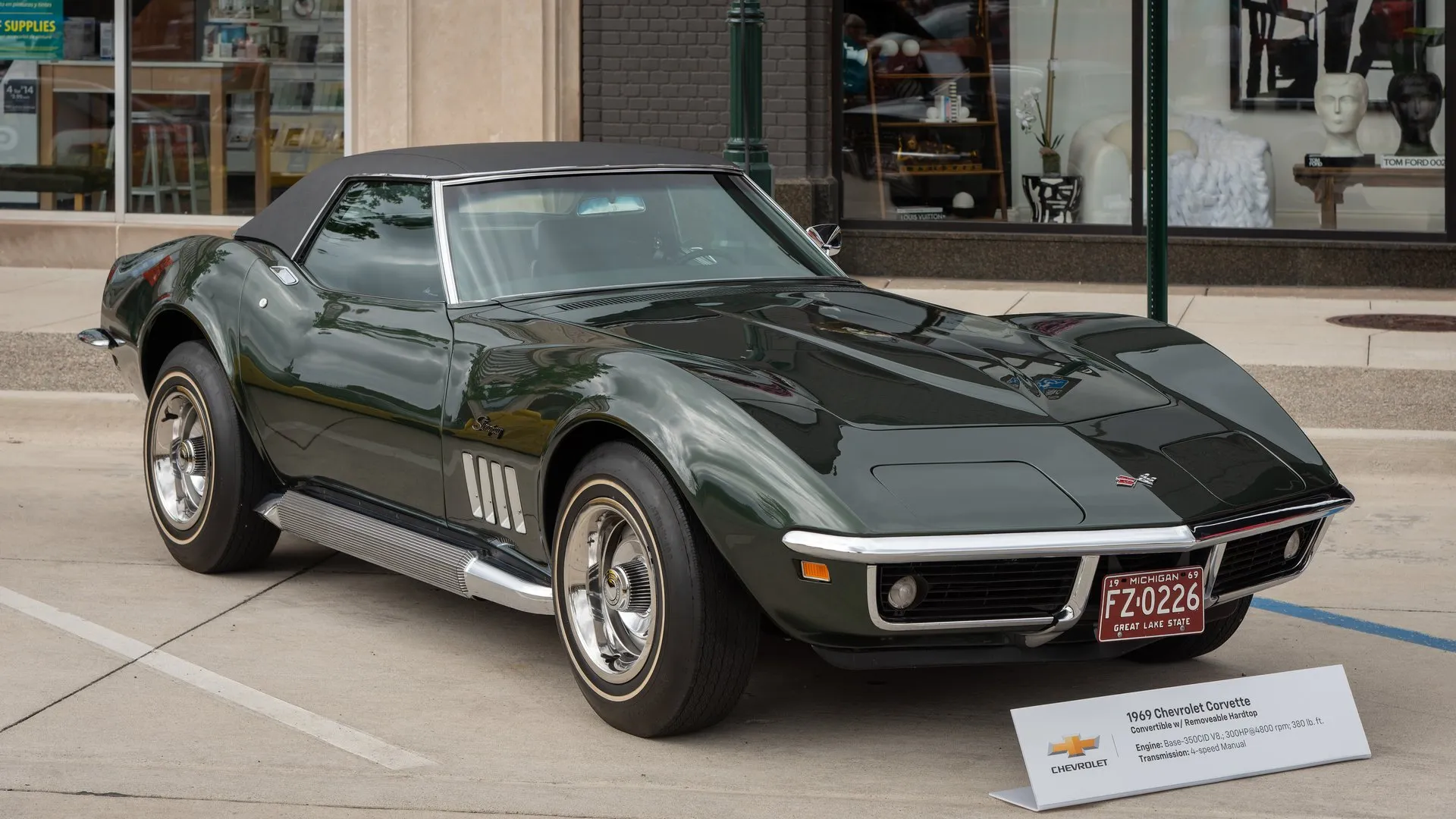 A dark green 1969 Corvette convertible with a black removable hardtop and side exhaust pipes below the doors, parked at a curb beside a display card, seen from the front right.