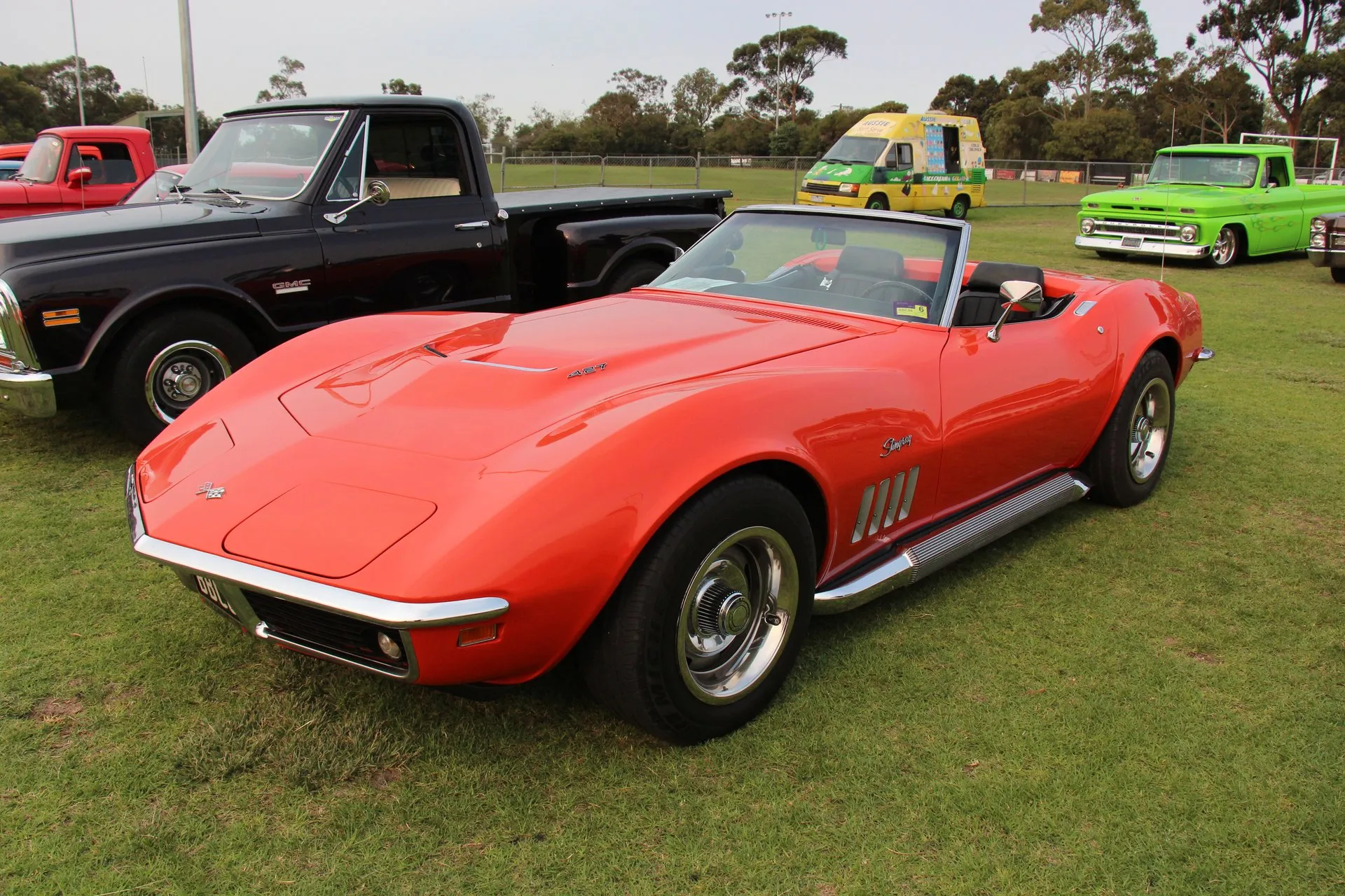 A red 1969 Corvette convertible with its top down, parked on grass, seen from the front left, with a 427 emblem on its raised hood and side exhaust pipes below the doors.