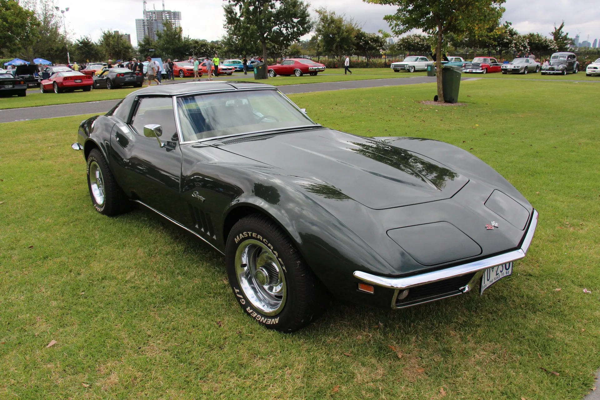 A dark-painted 1969 Corvette coupe parked on grass at a car show, seen from the front right, with Stingray script above four vertical slots on the front fender and white-letter tires.