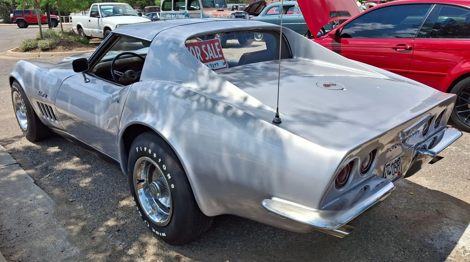 The rear of a silver 1969 Corvette coupe in a parking lot, seen from the rear left, with a For Sale sign in the rear window, four round taillights and a Corvette nameplate.