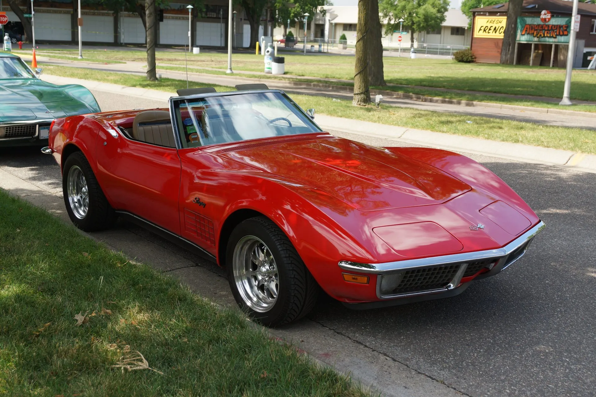 A red C3 Corvette convertible with its top down and a tan interior, parked at a curb and seen from the front right, with an egg-crate grille, Stingray script and an egg-crate louver on the front fender, flared wheel openings and polished aftermarket wheels.