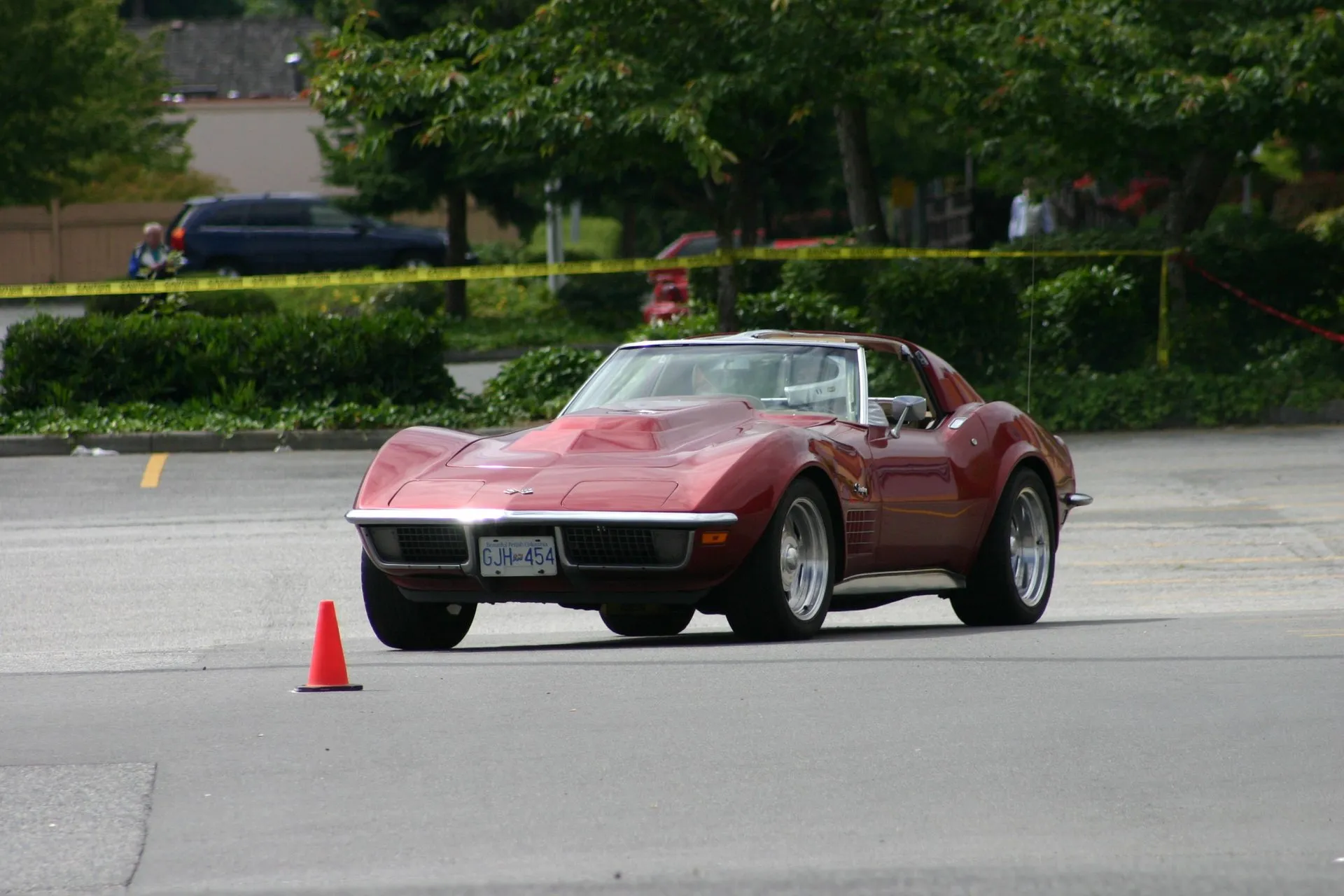 A maroon C3 Corvette coupe cornering on an asphalt autocross course past an orange traffic cone, seen from the front, with a domed hood, a helmeted driver and a British Columbia plate.
