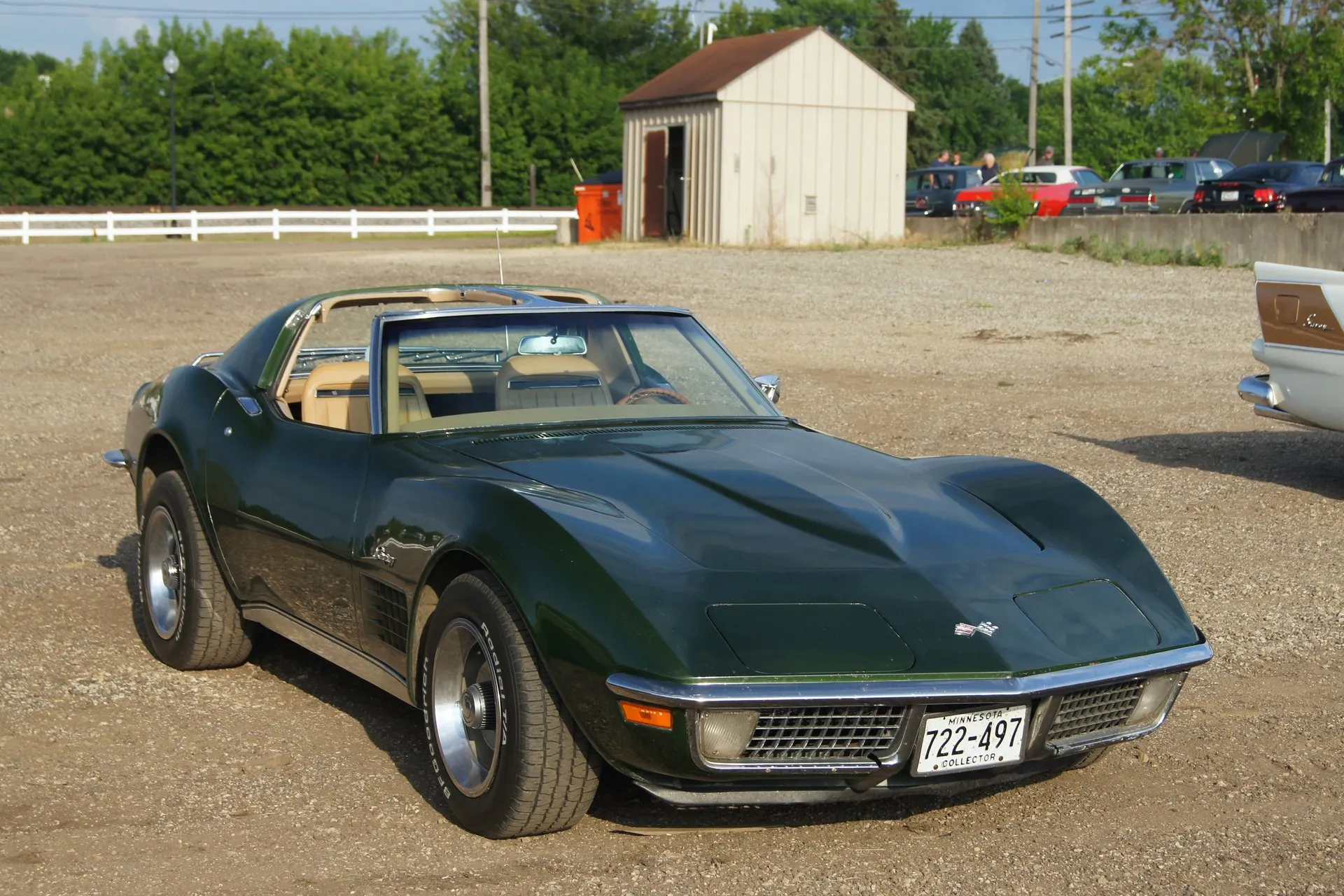 A dark green C3 Corvette coupe with its roof panels off and a tan interior, parked on gravel and seen from the front right, with an egg-crate grille, chrome bumpers, steel wheels with trim rings and center caps, and a Minnesota collector plate.