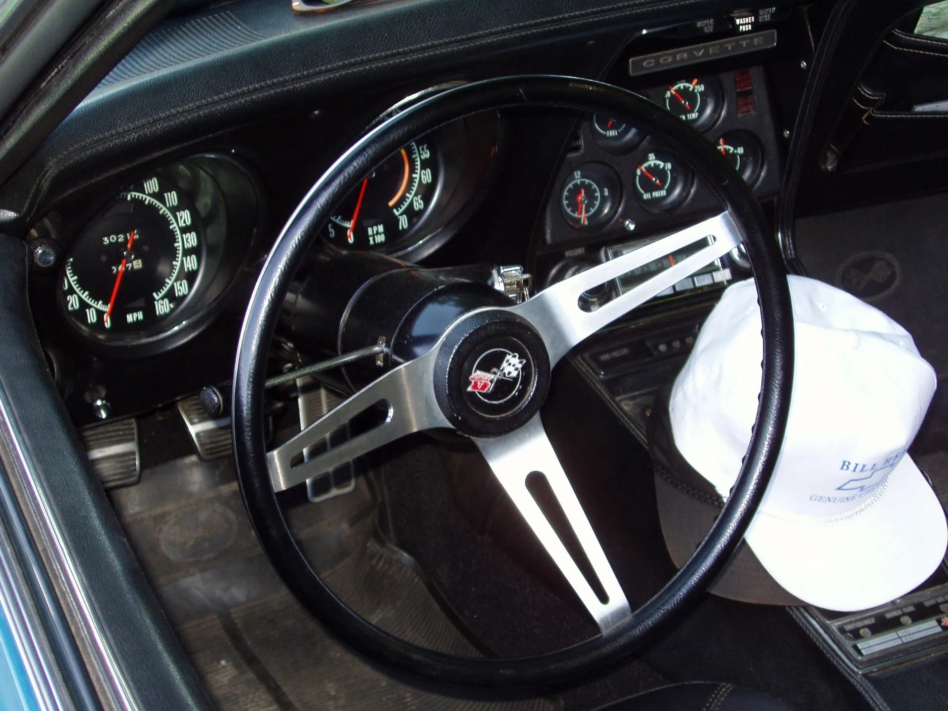 The driver's side of a C3 Corvette interior: a three-spoke steering wheel with a crossed-flags center, a large speedometer and tachometer behind it, small gauges and a radio in the center stack under a Corvette nameplate, and a white cap hung on the wheel's right side.