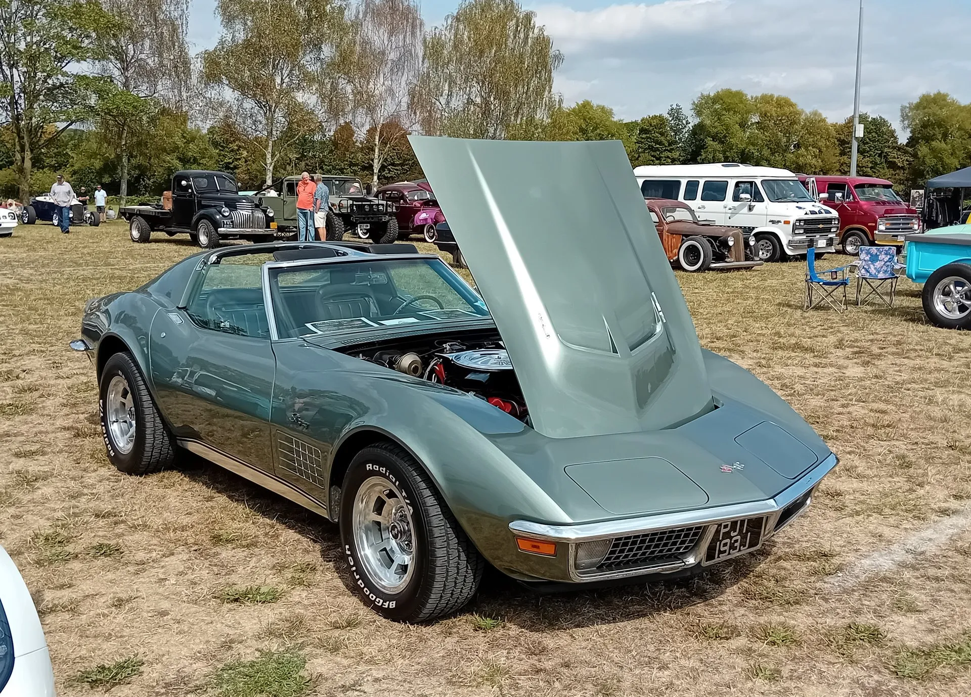 A gray-green C3 Corvette coupe with its hood raised, parked on dry grass at a car show and seen from the front right, with a chrome air cleaner visible in the engine bay and older trucks and vans behind it.