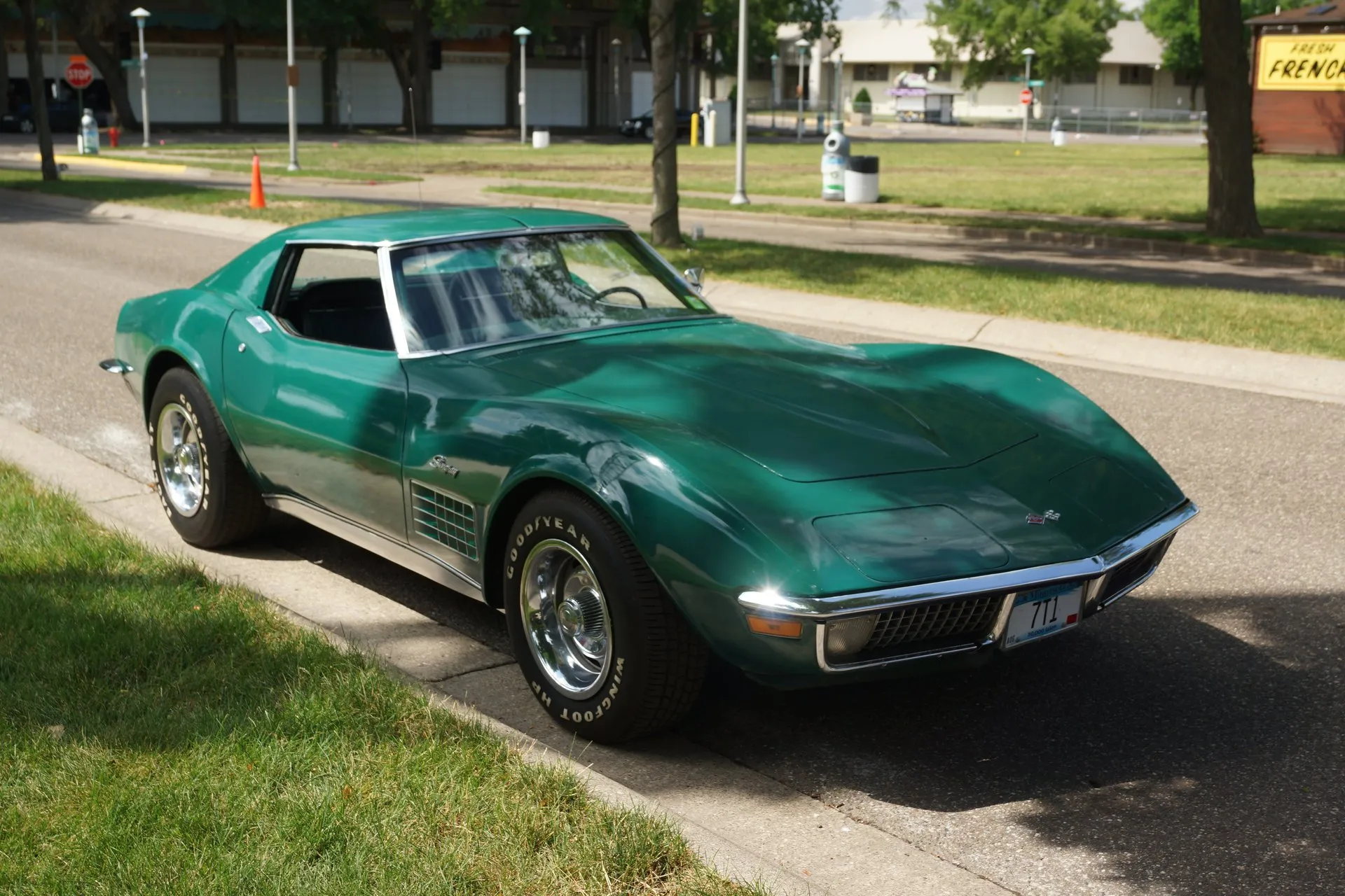 A dark green C3 Corvette coupe with its roof panels in, parked at a curb and seen from the front left, with a domed hood, chrome bumpers, an egg-crate grille and fender louver, and raised white-letter Goodyear tires on steel wheels with trim rings.