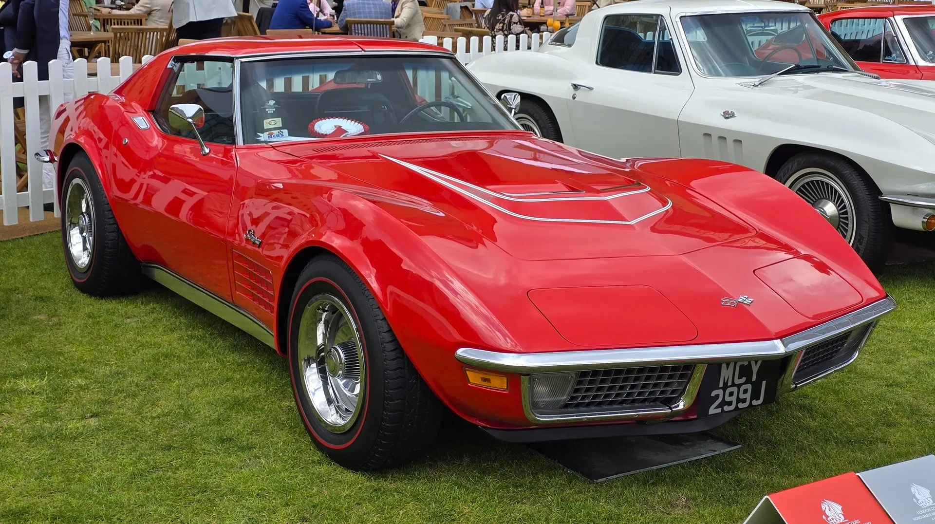 A red C3 Corvette coupe on grass at a show, seen from the front right, with a domed hood outlined in white stripes, chrome bumpers, red-line tires on chrome wheels and a British plate, beside a white earlier Corvette.