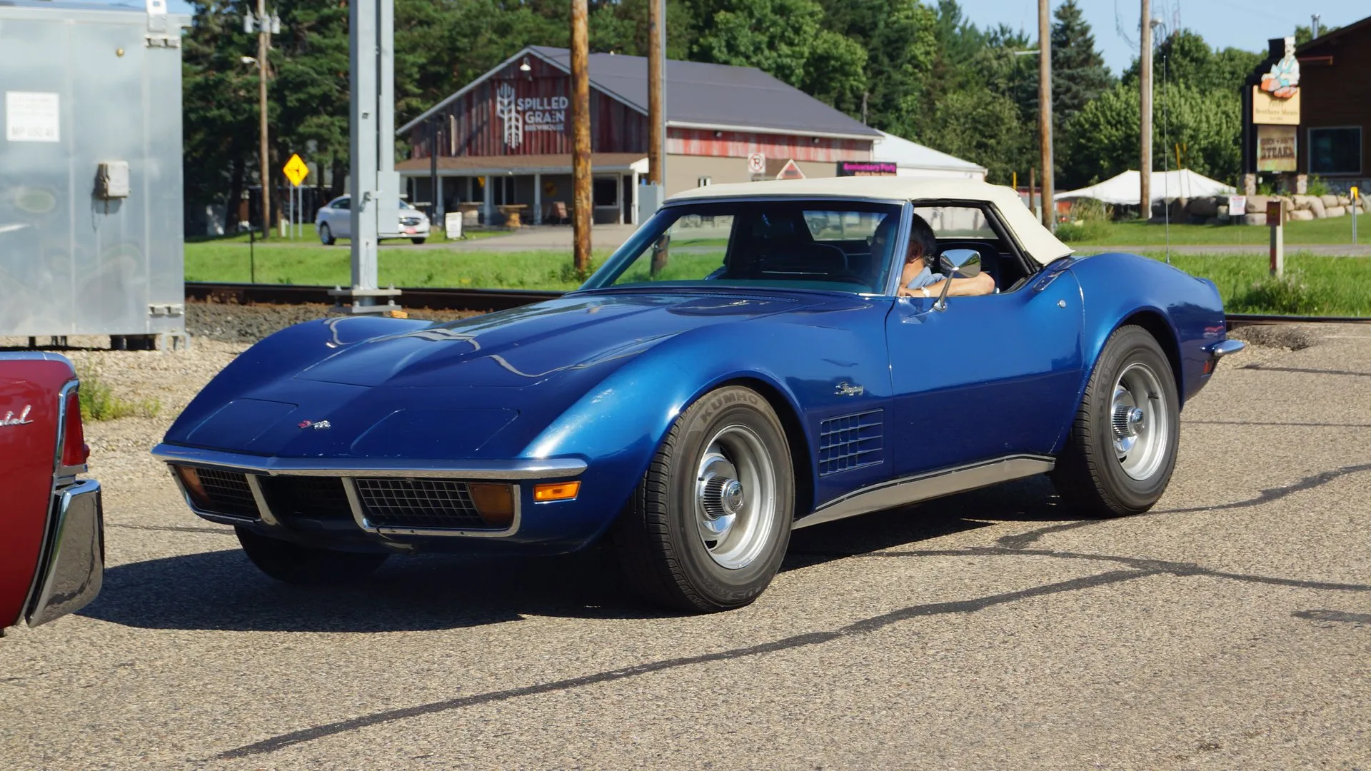 A metallic blue C3 Corvette convertible with a white soft top up, driving across a parking lot, seen from the front left, with an egg-crate grille, chrome front bumpers, amber parking lamps and a grille in the front fender vent.