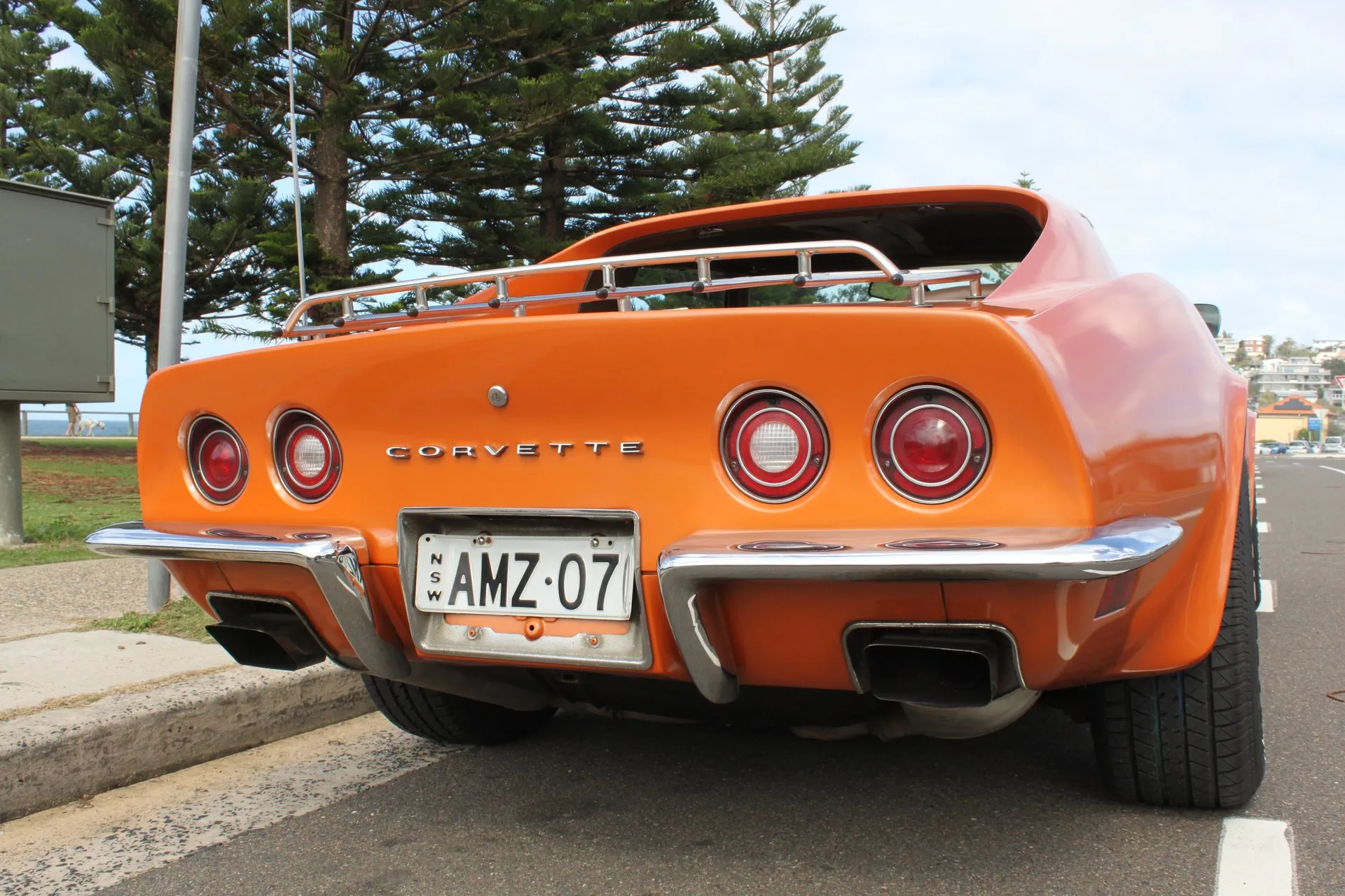 The rear of an orange C3 Corvette coupe parked at a curb, seen from low down, with four round taillights, Corvette lettering, a two-piece chrome rear bumper, rectangular exhaust outlets and a chrome luggage rack on the deck.