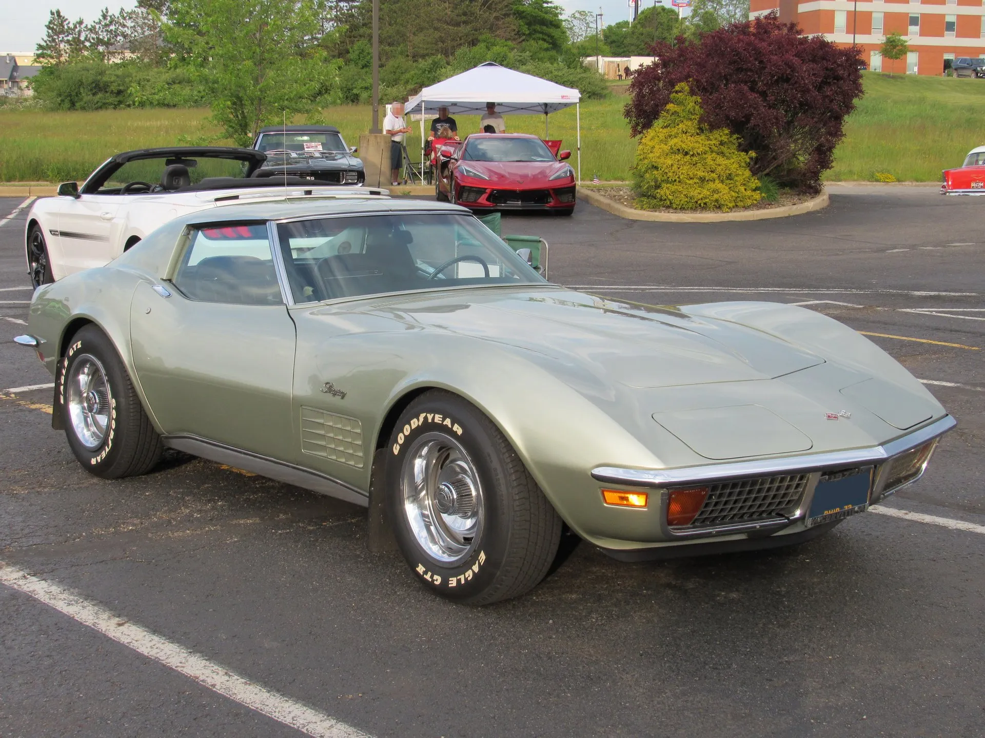 A pale silver-green C3 Corvette coupe in a parking lot, seen from the front right, with chrome bumpers, an egg-crate grille, a fender grille under the Stingray script, Rally wheels and white-letter Goodyear tires.