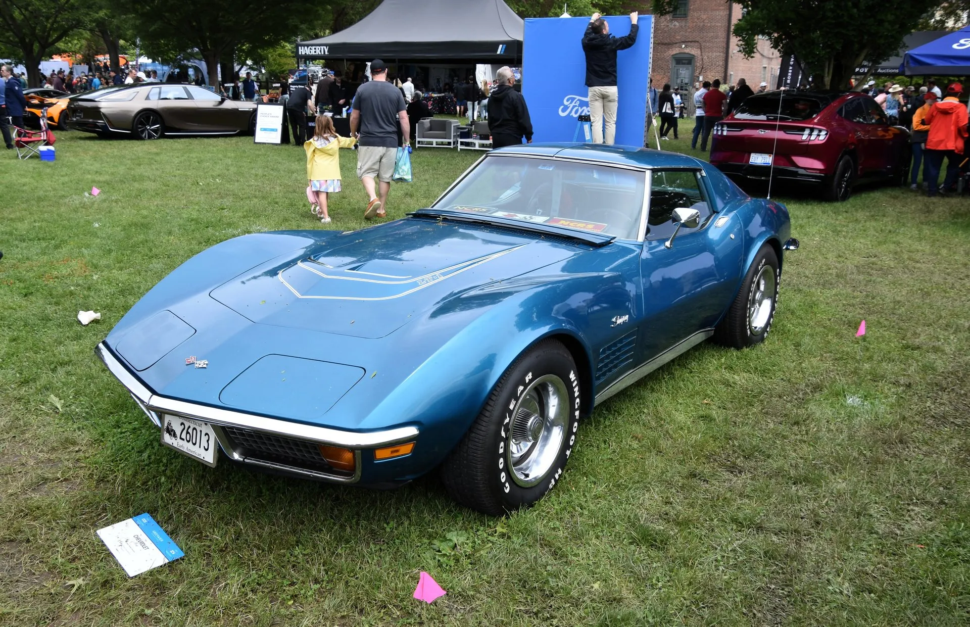 A blue C3 Corvette coupe on the grass at a car show, seen from the front left, with LT-1 stripes on its domed hood, chrome bumpers, amber parking lamps, Stingray script, a grille in the fender vent and white-letter Goodyear tires.