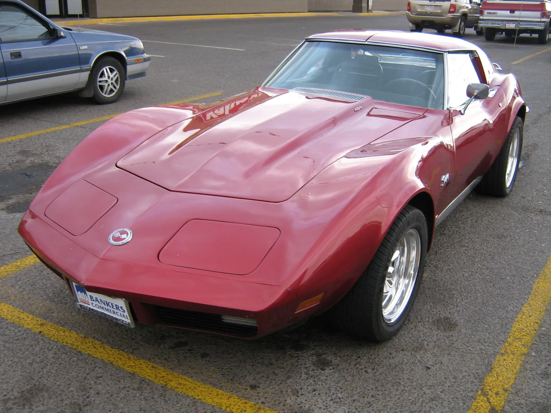 The same red Corvette coupe from the front left, with a smooth body-color nose, closed headlight doors, a raised hood with a grille strip at its rear edge, a small badge on the hood and an advertising plate on the front.