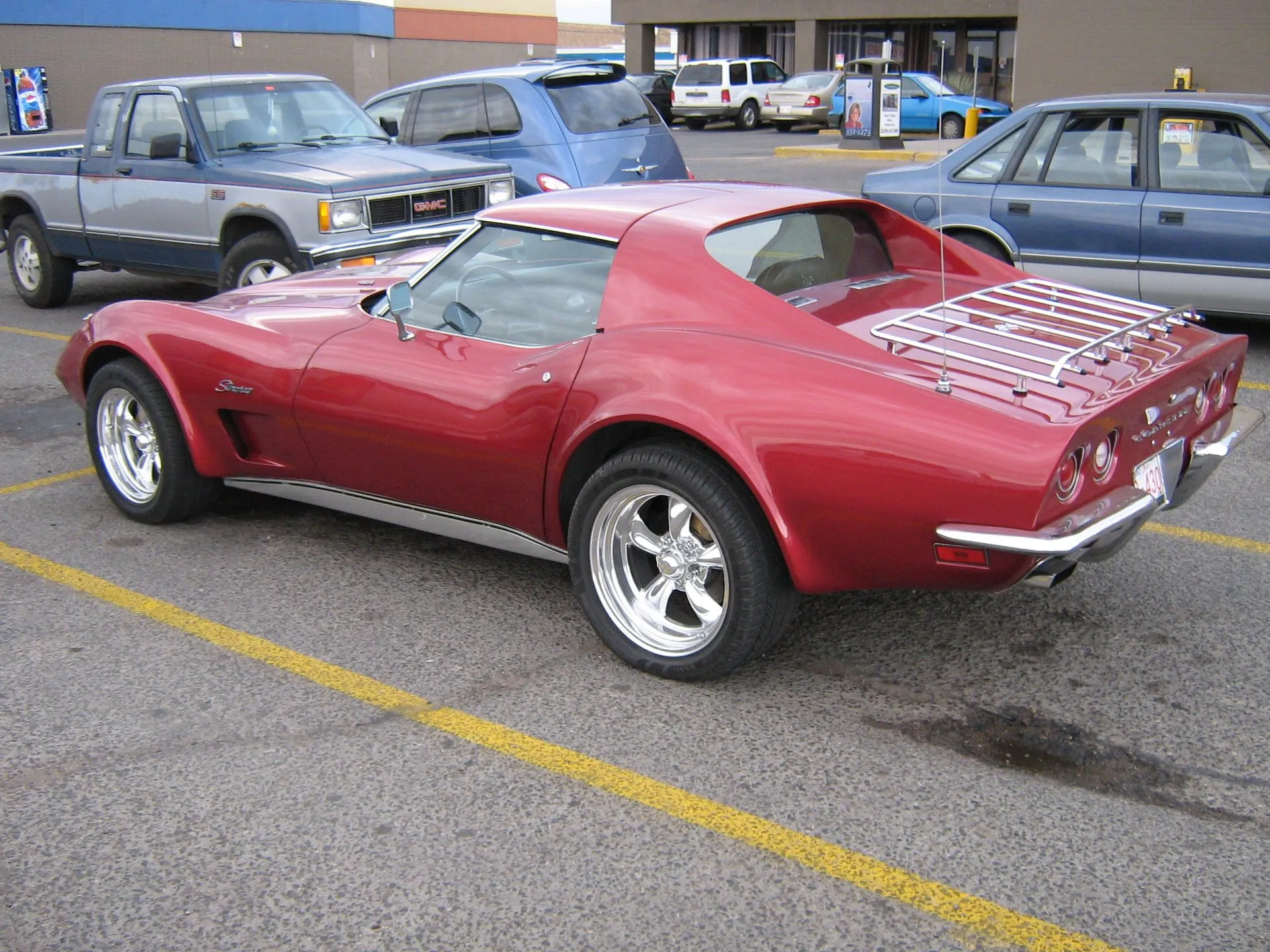 A red C3 Corvette coupe in a parking lot, seen from the rear left, with a chrome rear bumper, four round taillights, a chrome luggage rack on the rear deck, Stingray script above an open fender vent and polished five-spoke wheels.