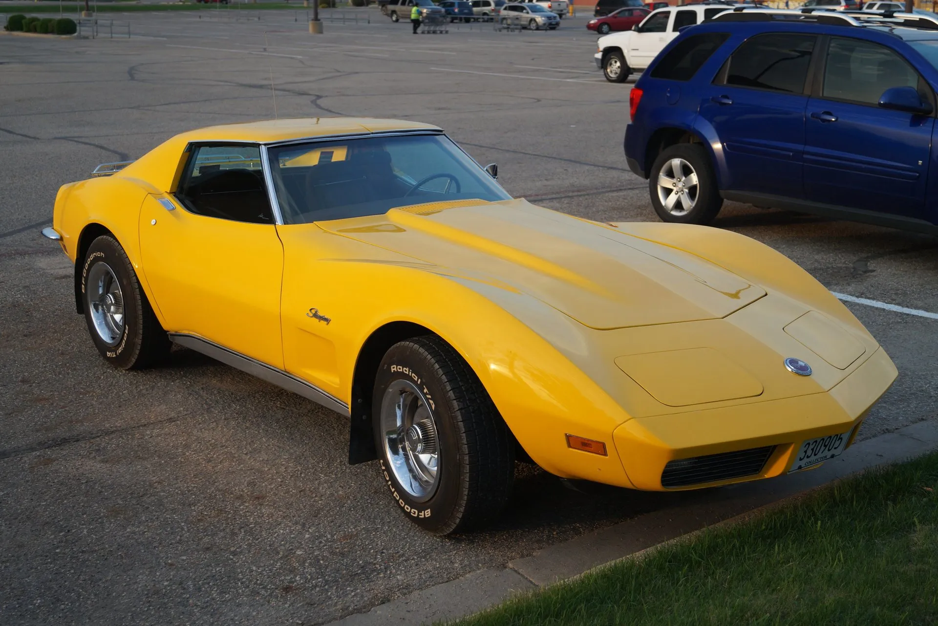 A yellow C3 Corvette coupe parked in a lot, seen from the front right, with a body-color nose, Stingray script on the front fender, steel wheels with chrome trim rings and center caps, and white-letter radial tires.