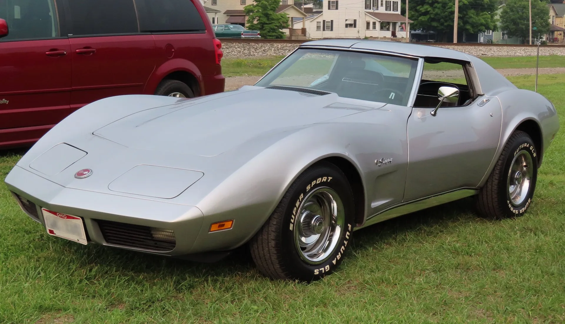 A silver C3 Corvette coupe parked on grass, seen from the front left, with a smooth body-color nose, closed headlight doors, Stingray script on the front fender and white-letter tires.