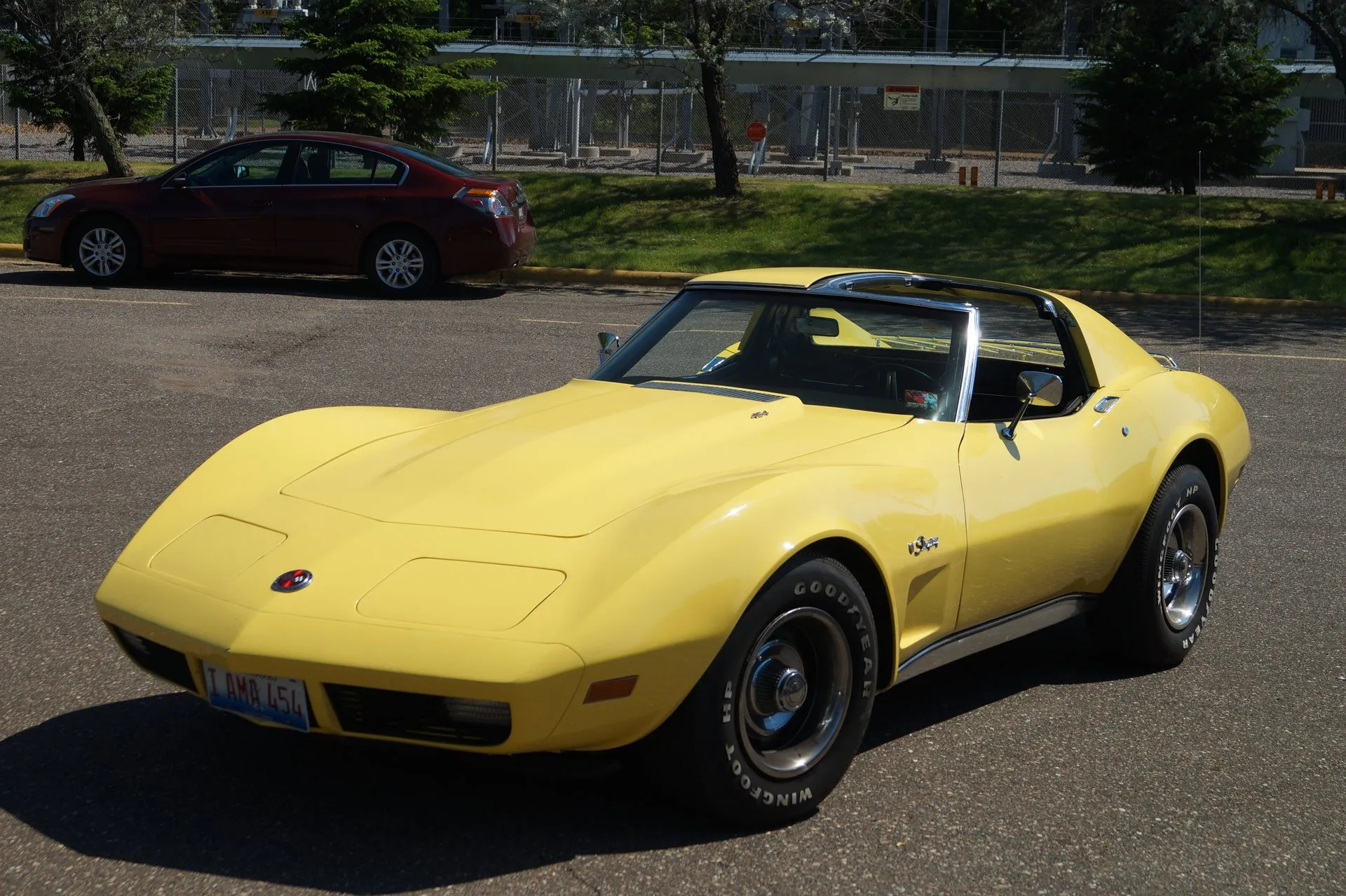 A yellow C3 Corvette coupe in a parking lot, seen from the front left, with a small badge on the hood, an emblem on the front fender, white-letter tires and a front plate reading I AMA 454.