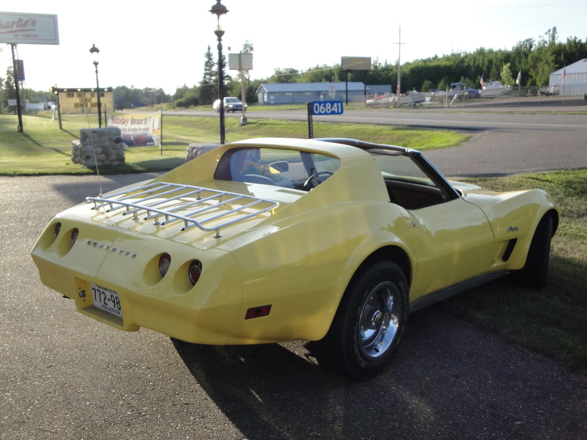 A yellow C3 Corvette coupe parked at a roadside, seen from the rear left, with a chrome luggage rack on the deck, Corvette letters between four round taillights and a body-color rear bumper with a seam down its middle.