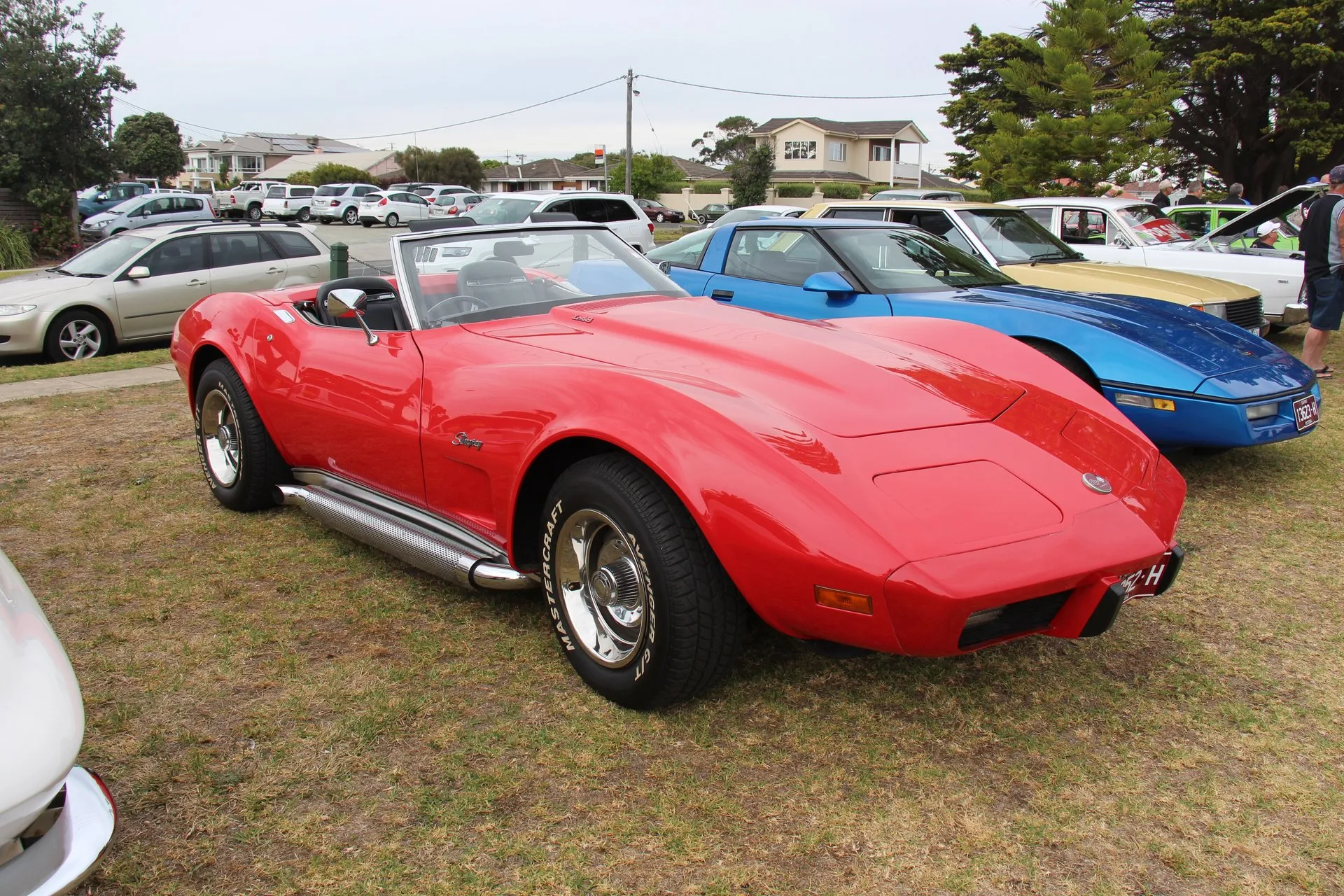 A red C3 Corvette convertible with its top down, parked on grass at a car show and seen from the front right, with Stingray script on the front fender, chrome perforated side exhaust pipes, chrome wheels with center caps and a small badge on the hood.