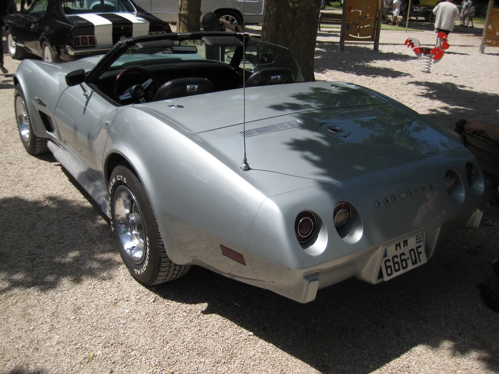 A silver C3 Corvette convertible with its top down, parked on gravel and seen from the rear left, with block letters spelling Corvette between four round taillights, two slotted vents on the rear deck, a smooth body-color rear bumper and Stingray script on the front fender.