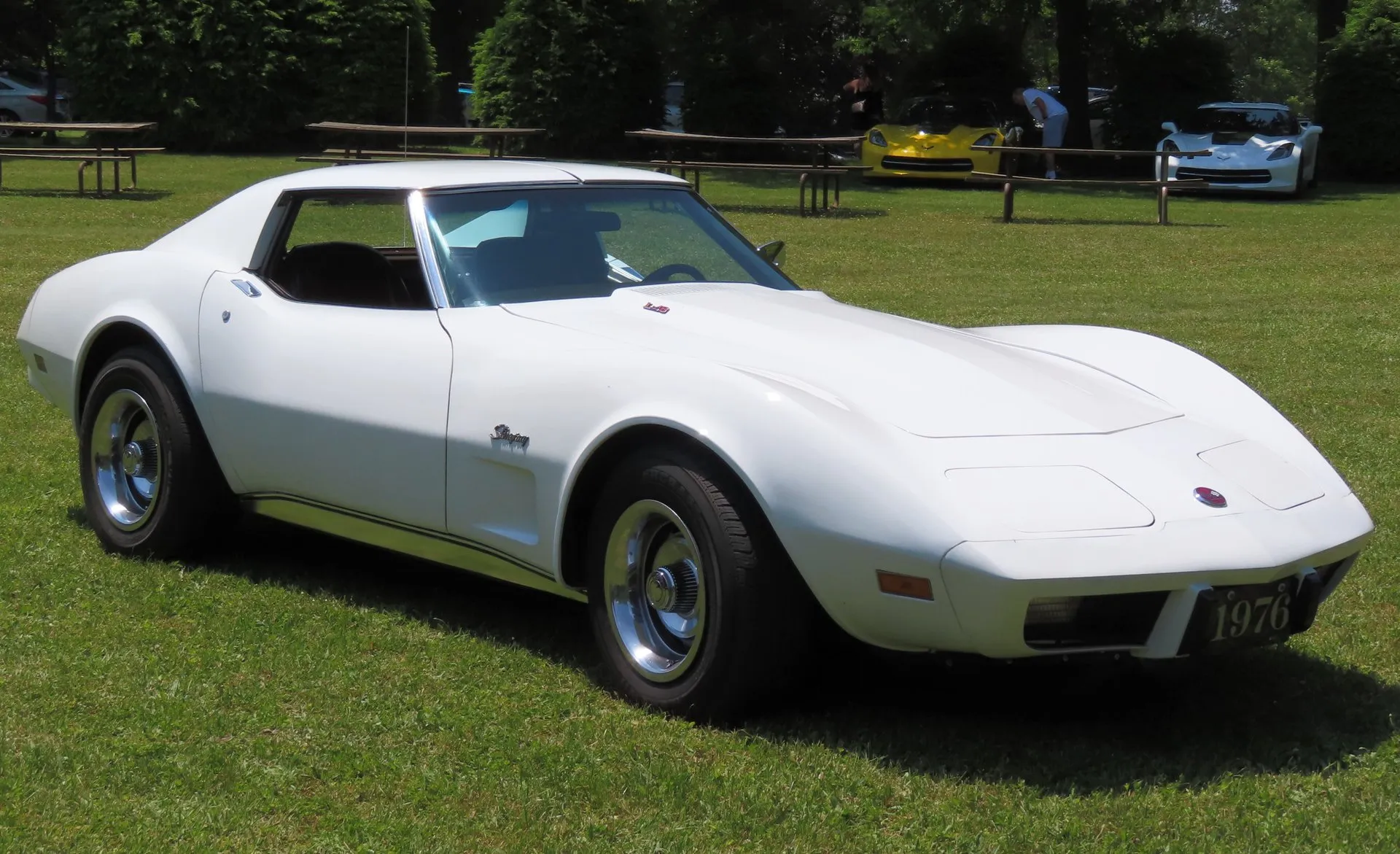 A white C3 Corvette coupe parked on grass at a car show, seen from the front and side, with Stingray script on the front fender, steel wheels with chrome trim rings and center caps, a small badge on the hood and a front plate frame reading 1976.