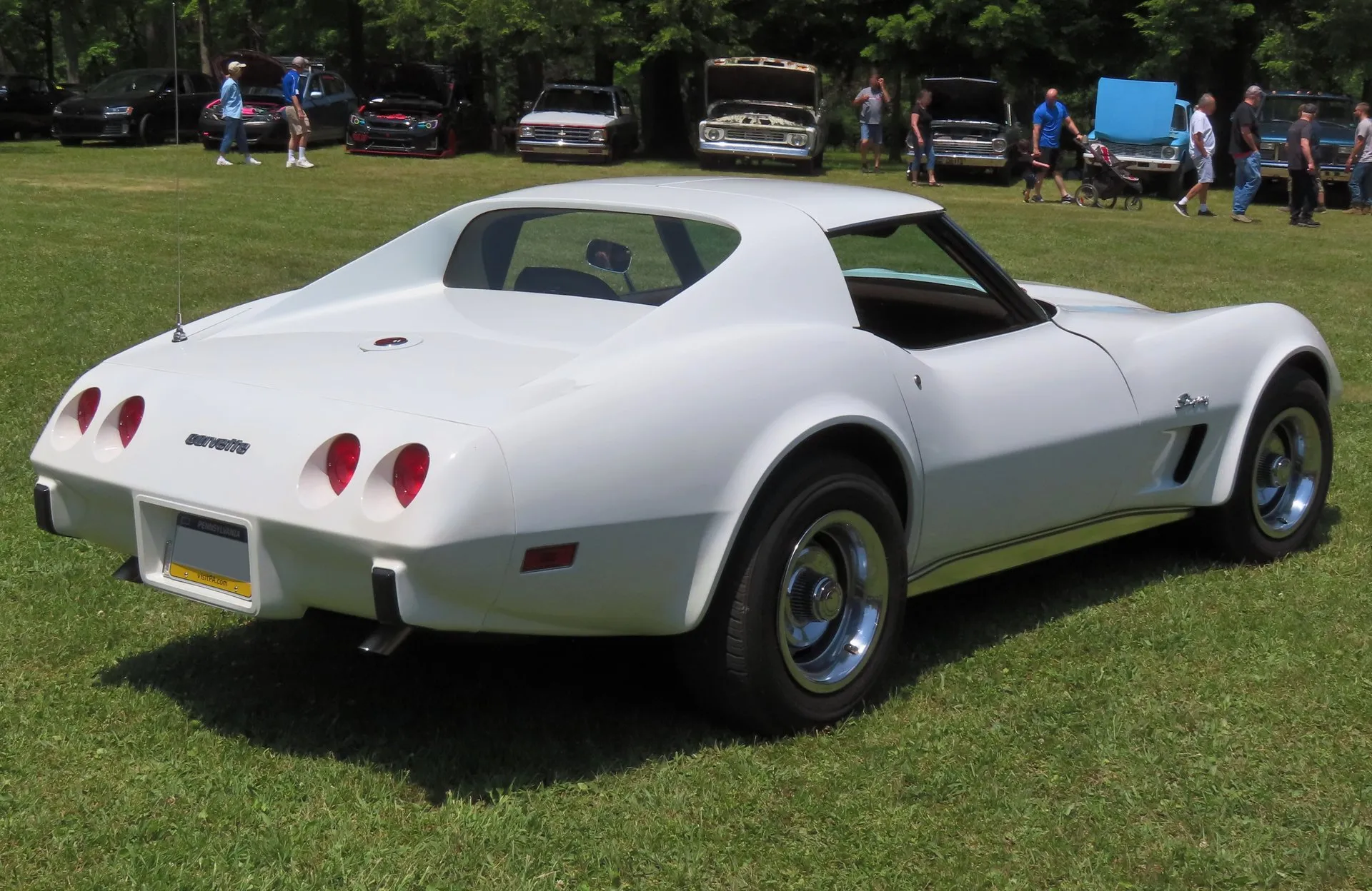 The rear of the same white Corvette coupe, seen from behind and to one side on grass, with a smooth deck behind the rear window, a one-piece Corvette script between four round taillights, a body-color rear bumper and Stingray script on the front fender.