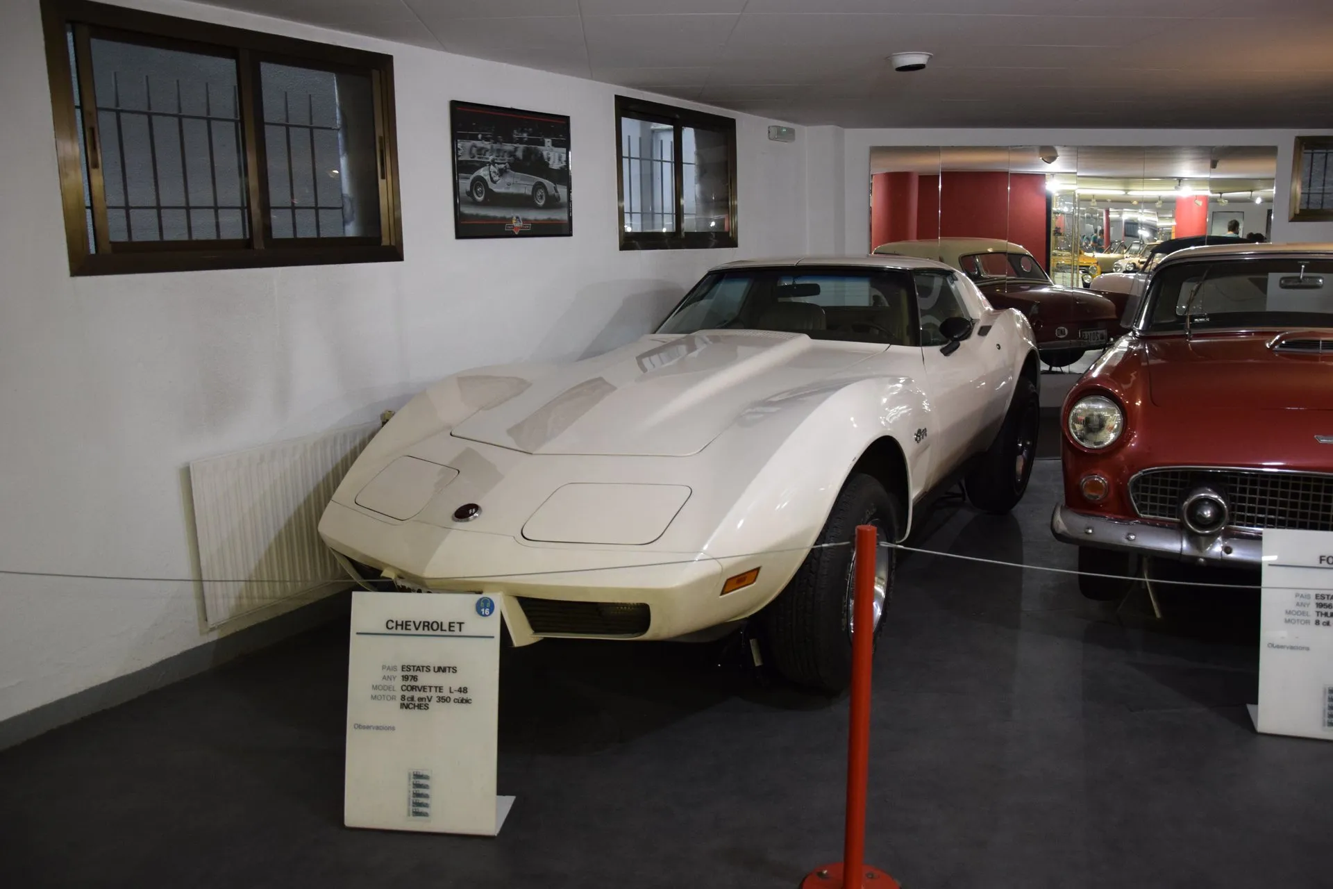 A cream-white C3 Corvette coupe on display behind a rope in a museum, seen from the front left, beside a red 1950s car, with a placard in front reading 1976, Corvette L-48, 8 cylinders, 350 cubic inches.