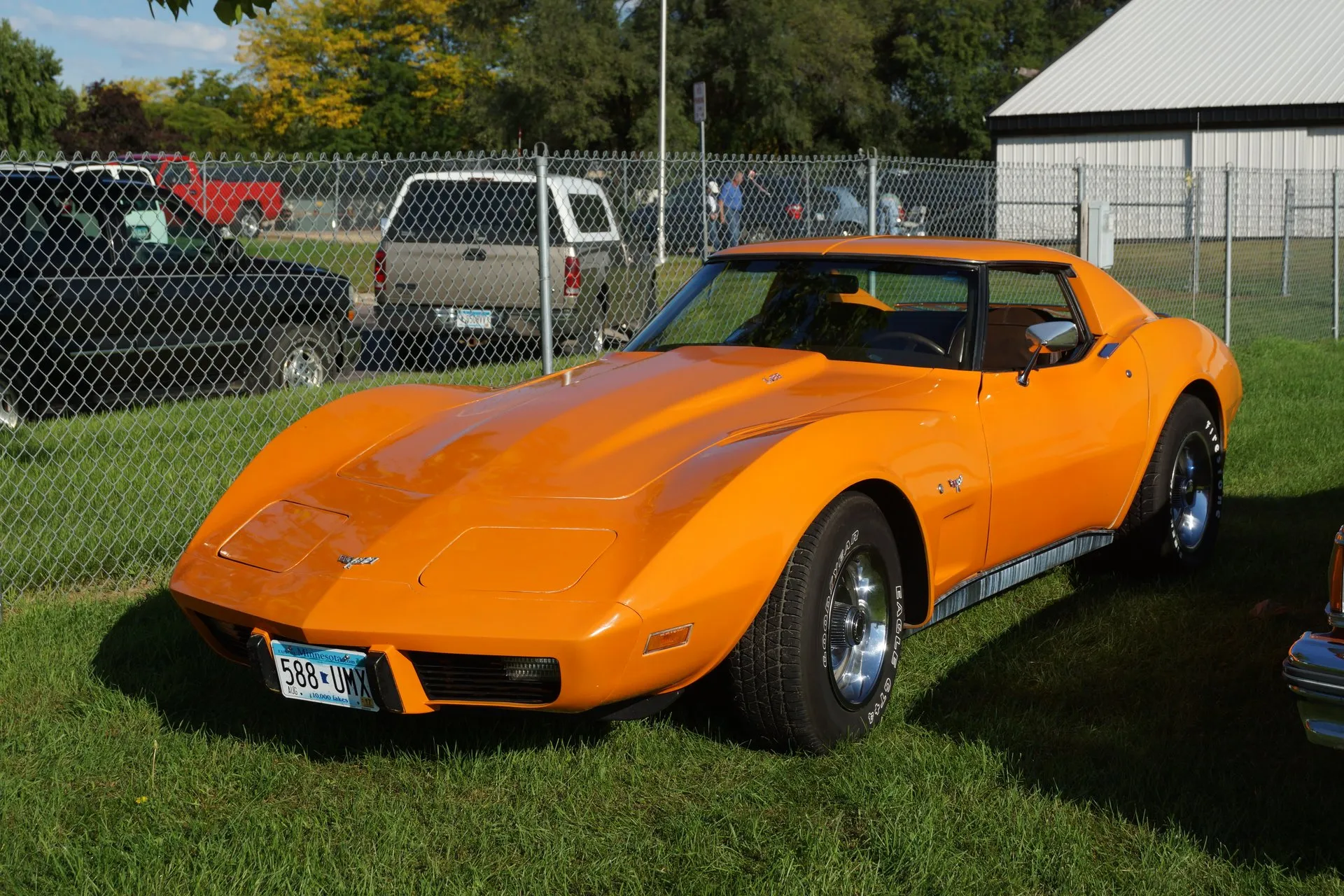 An orange C3 Corvette coupe parked on grass beside a chain-link fence, seen from the front left, with crossed flags on the nose and fender, a small badge on the hood, aluminum wheels and white-letter tires.