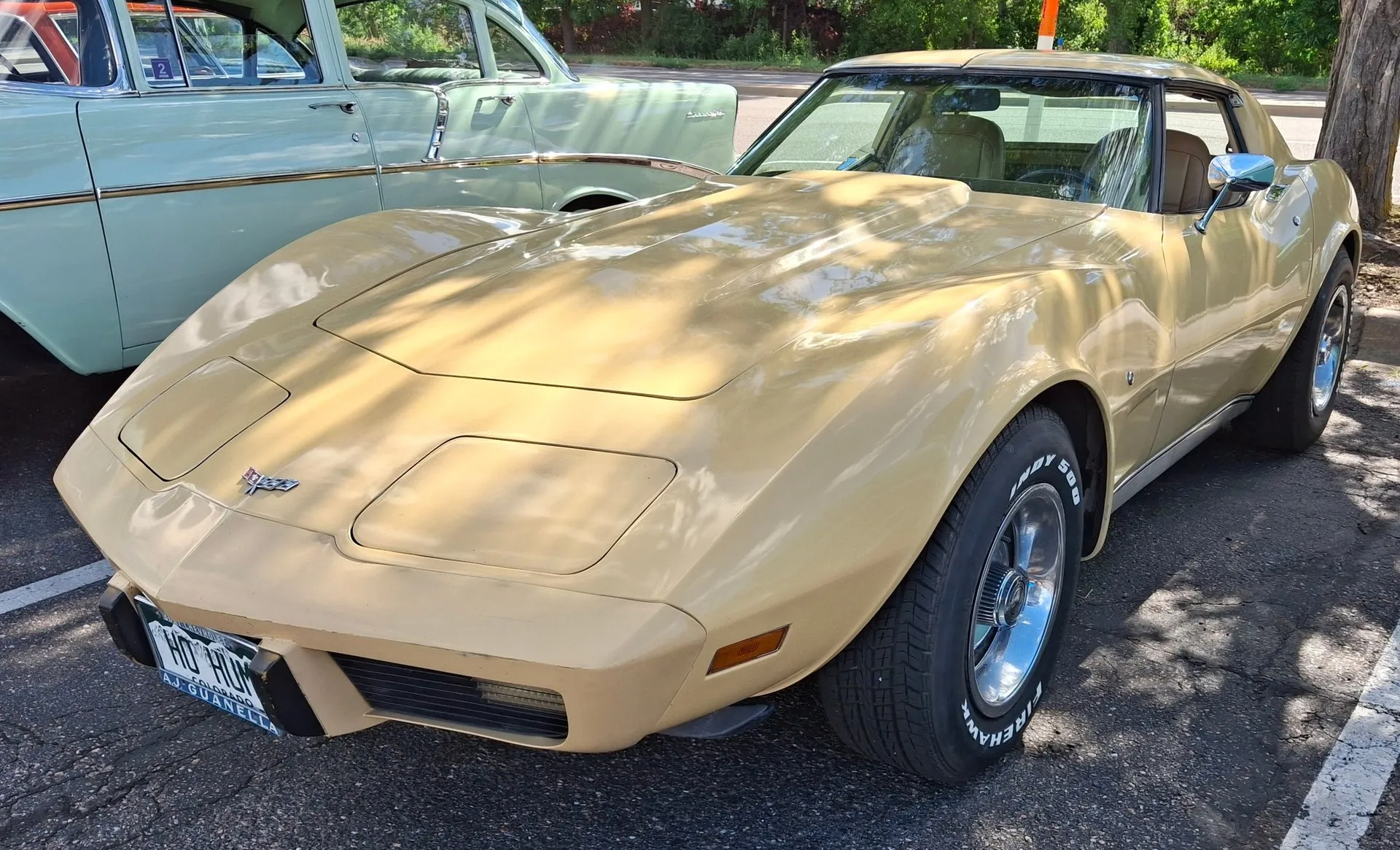 A tan C3 Corvette coupe parked at a car show, seen from the front left, with a crossed-flags emblem on the nose, a small round lock cylinder on the left front fender behind the wheel, no fender emblem or script, a black windshield surround and chrome wheels with white-letter tires.