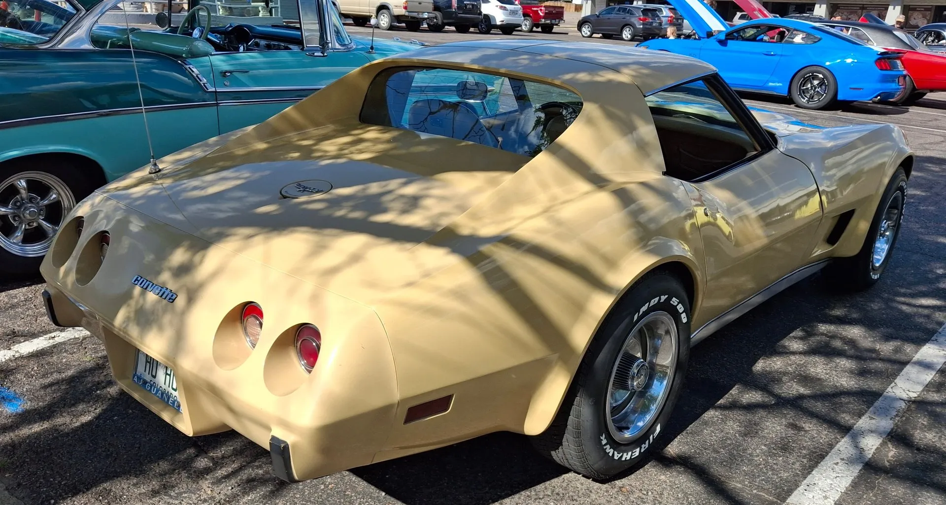 The rear of the same tan Corvette coupe from the right rear corner, showing the upright rear window set between the roof's flying buttresses, a smooth deck, a crossed-flags emblem on the fuel filler door and Corvette script between four round taillights.