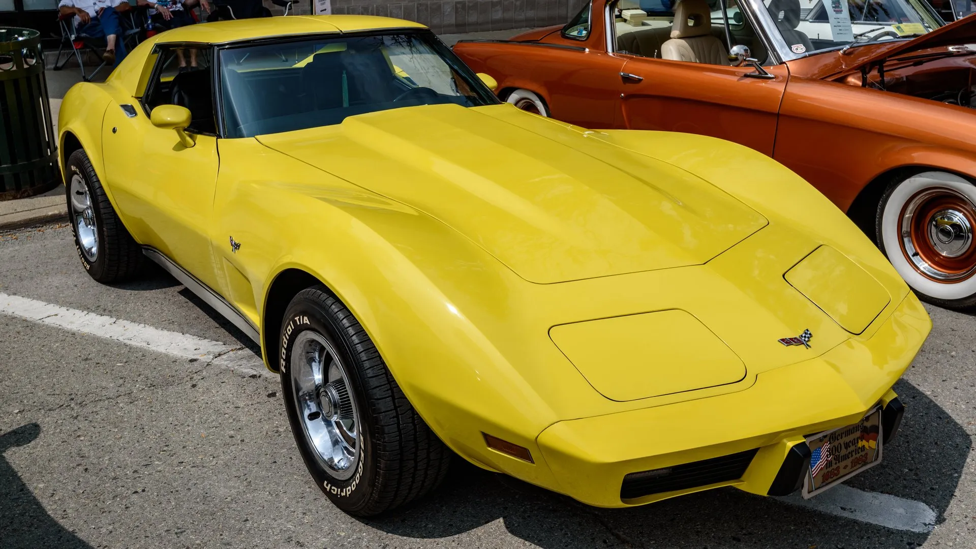 A yellow C3 Corvette coupe parked on a street at a car show, seen from the front right, with crossed flags on the nose and front fender and aluminum wheels.